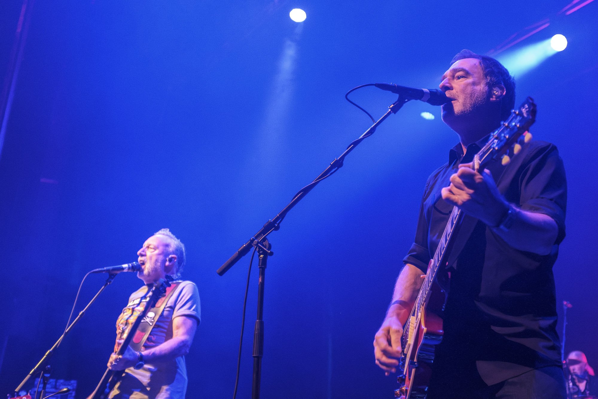 Two male musicians performing on stage with blue and purple lighting, one singing into a microphone and playing guitar, the other singing and playing guitar.