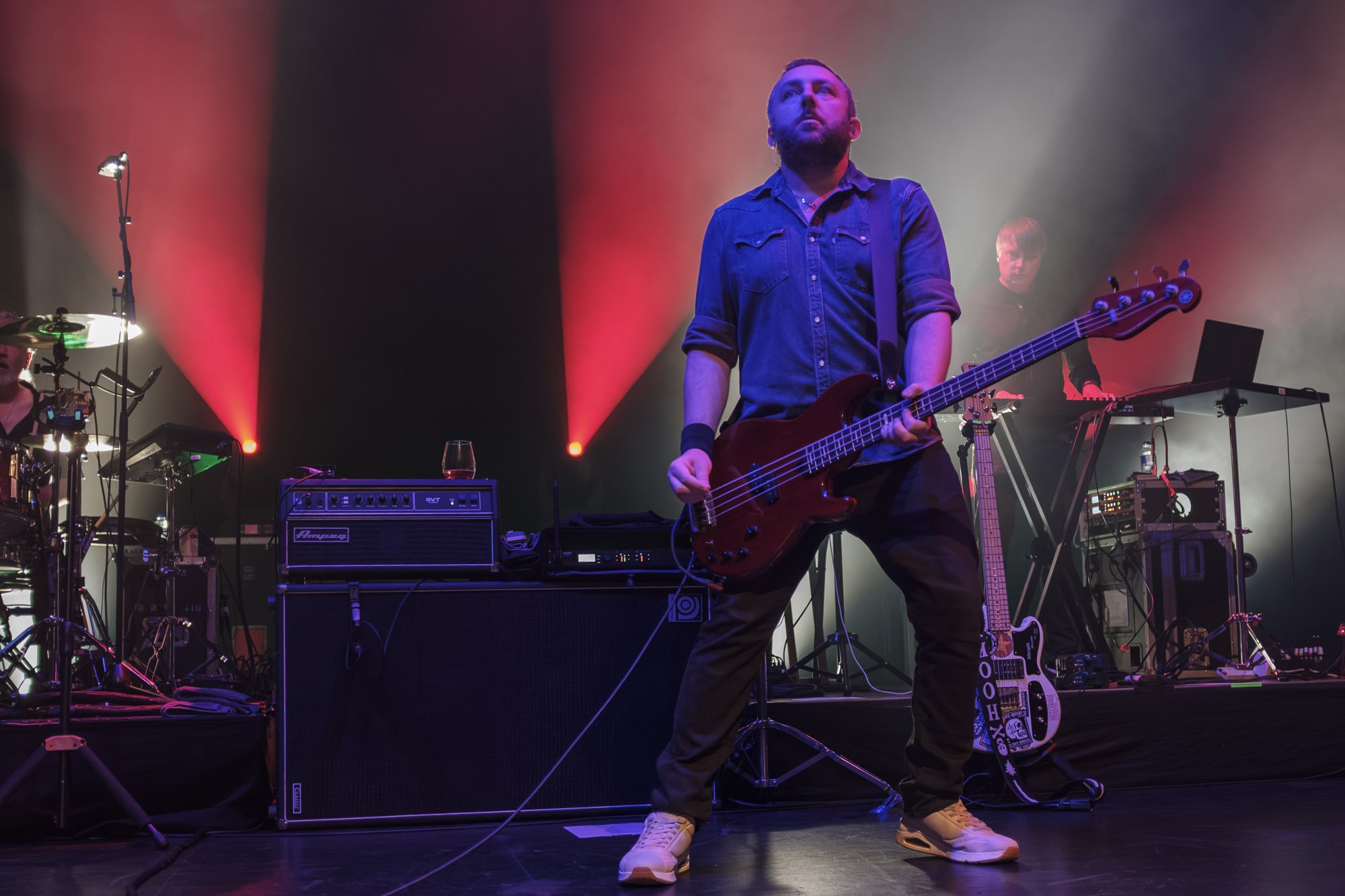 Musician standing on stage playing bass guitar with colorful stage lighting and audio equipment around.