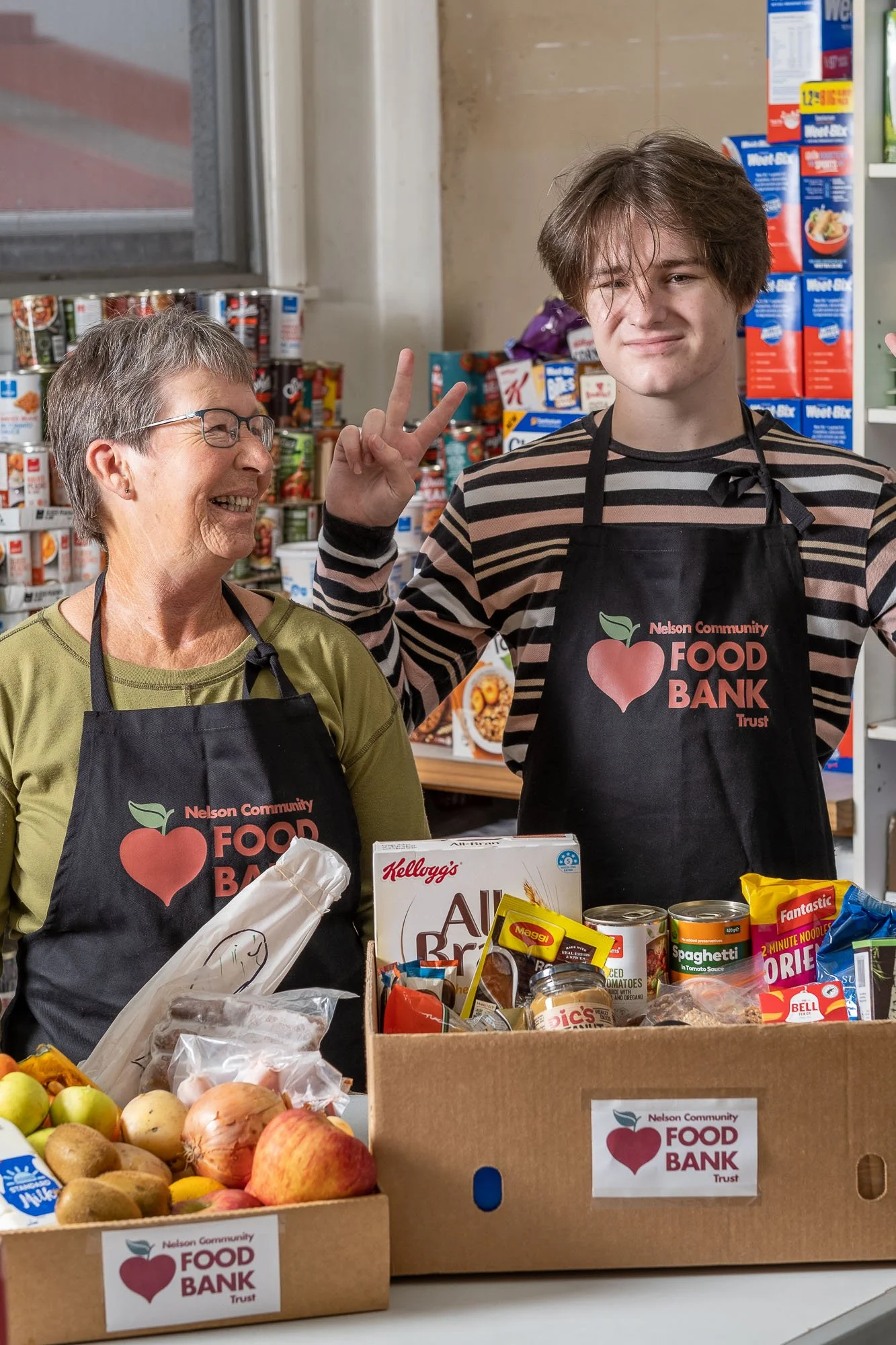 Two volunteers, an elderly woman and a young man, wearing Nelson Community Food Bank aprons, standing behind boxes of food in a food bank. The young man is making a peace sign with his fingers.