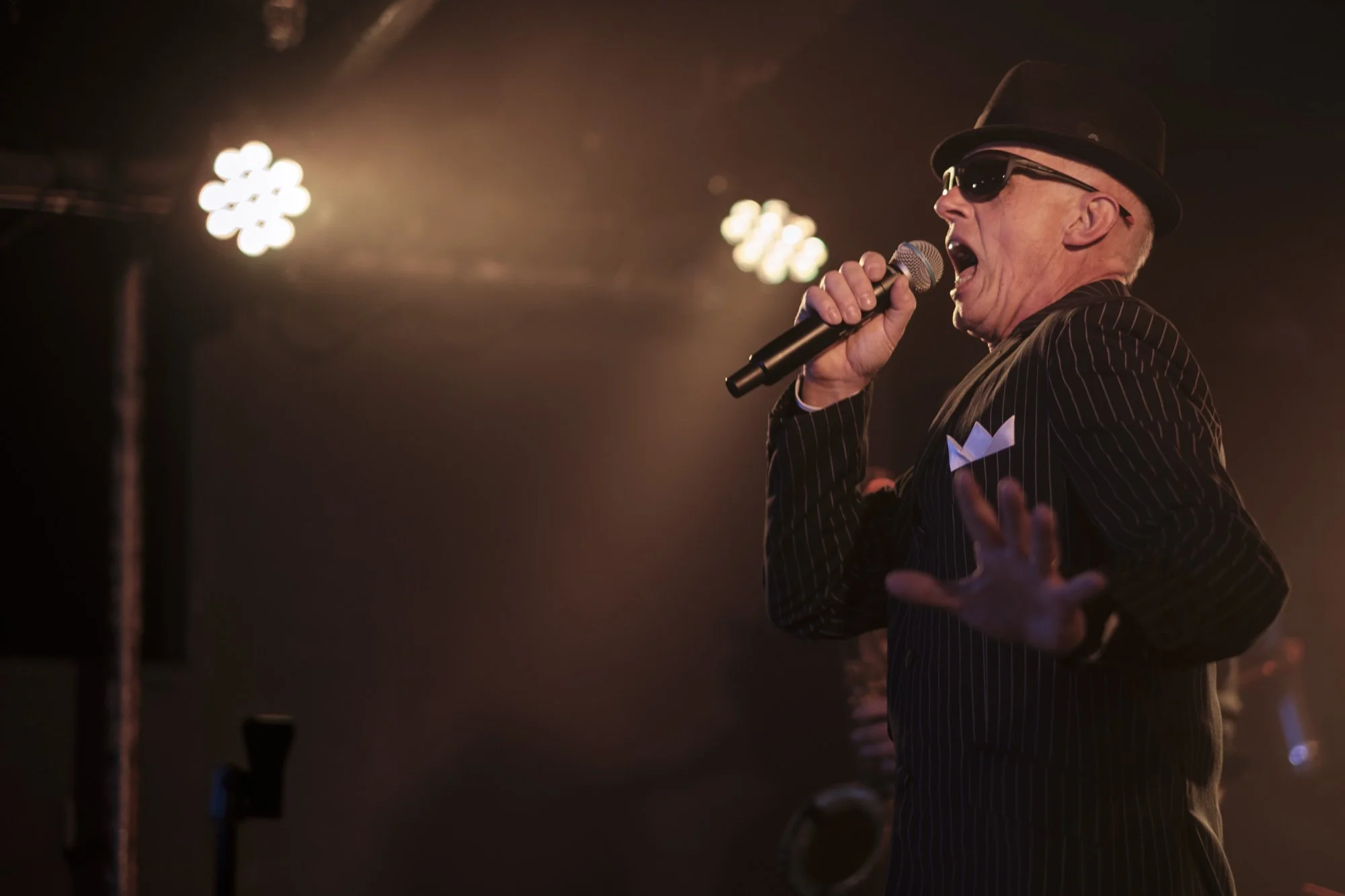 An older man dressed in a pinstripe suit, sunglasses, and a fedora hat singing or speaking into a microphone on stage with bright stage lights shining in the background.
