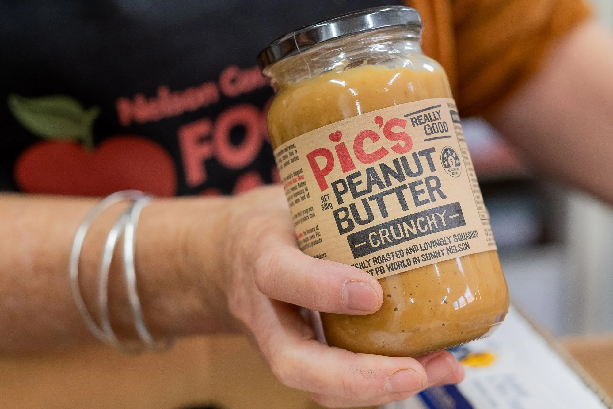 A person holding a jar of Pic's Really Good Peanut Butter Crunchy. The jar's label features red and black text on a tan background, and the person's arm is adorned with silver bangles.