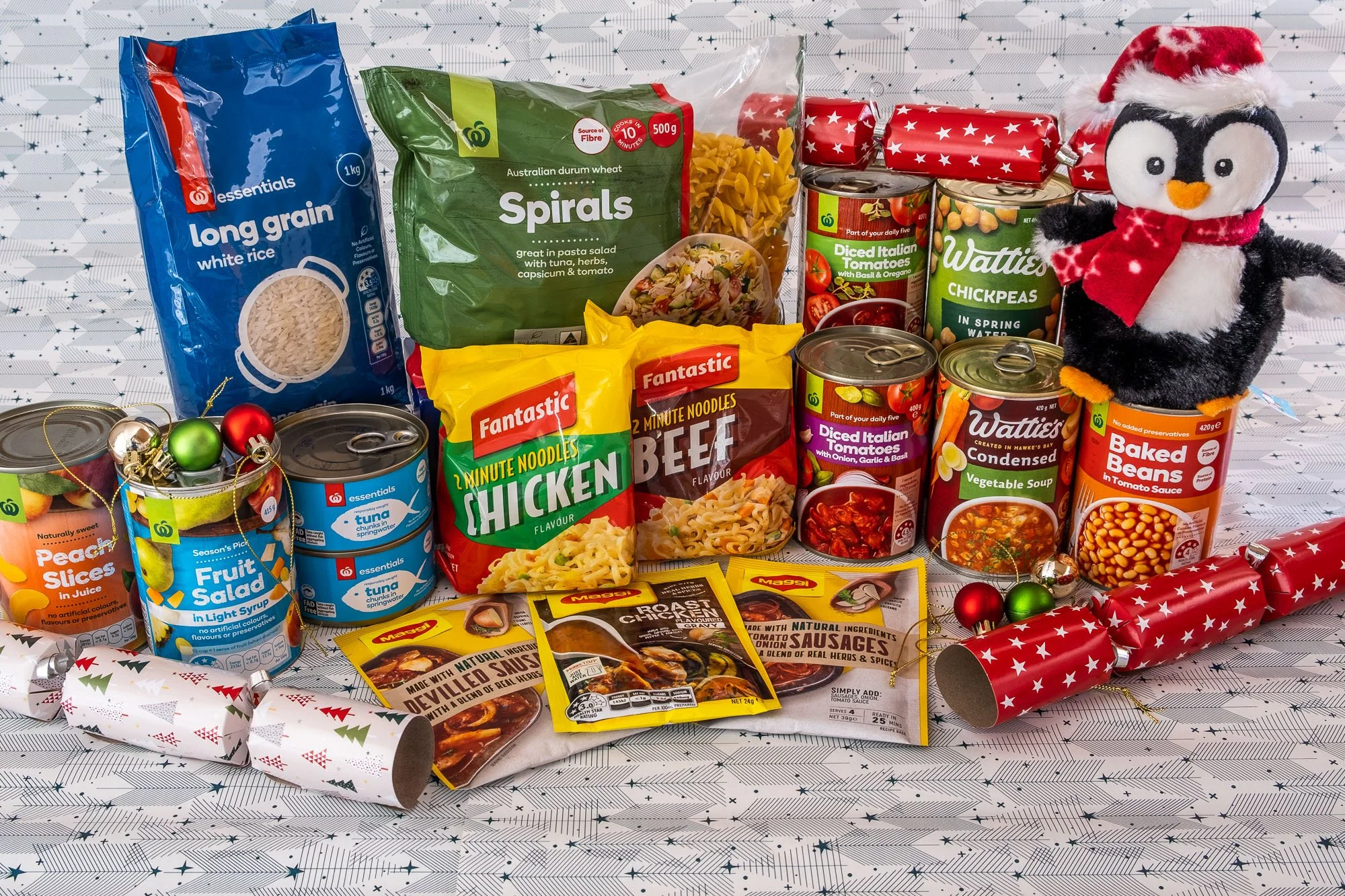 Assorted canned foods, dried pasta, snack noodles, sausage packets, festive party noise makers, a plush penguin toy wearing a Santa hat and scarf, and Christmas ornaments on a holiday-themed tablecloth.