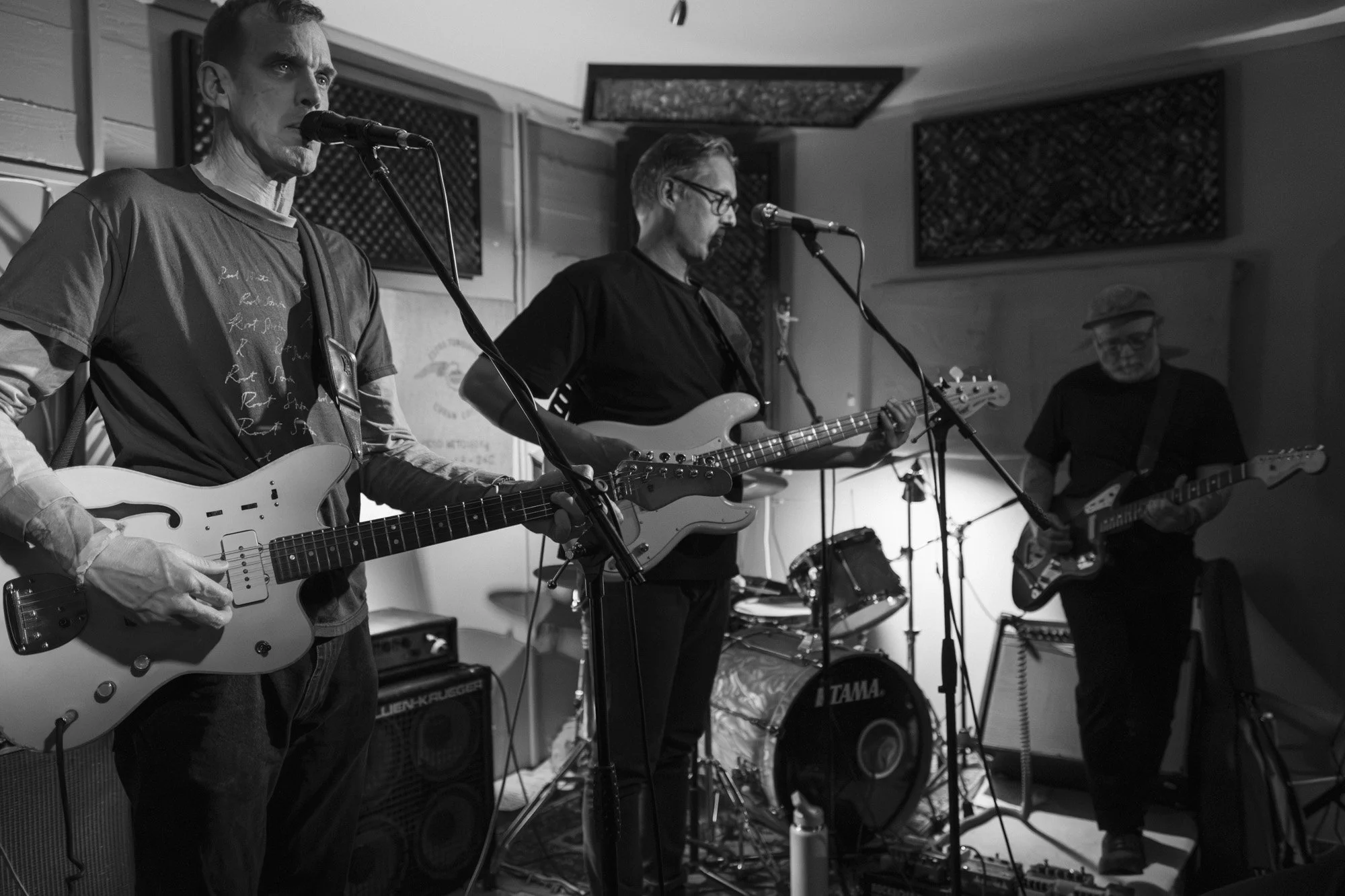 Three men playing electric guitars and singing into microphones during a band practice or performance in a room with acoustic wall panels. One man on the left is wearing a t-shirt and playing a white guitar, the man in the middle is wearing glasses a