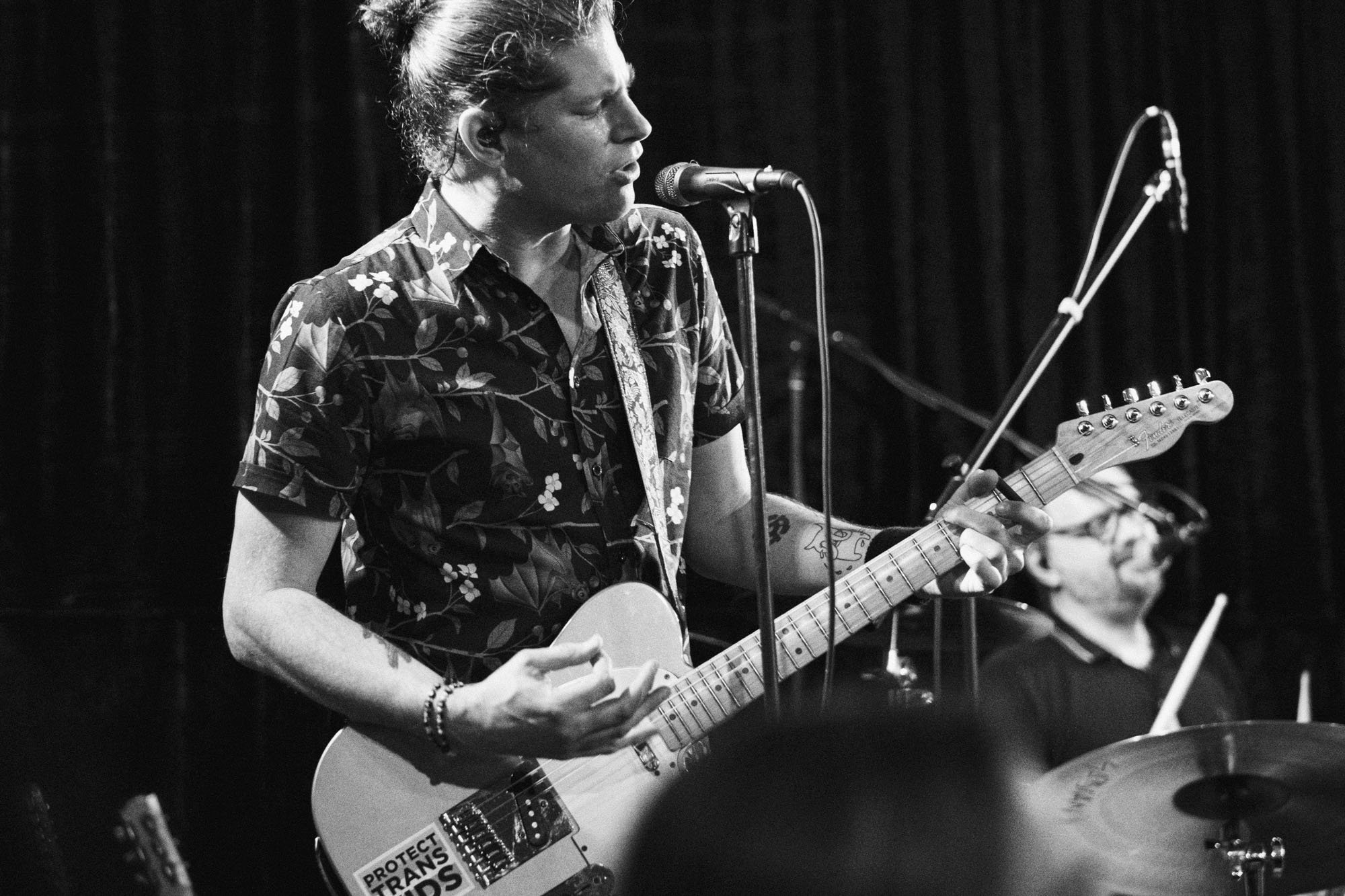 A black and white photo of a young man with hair tied back, playing an electric guitar and singing into a microphone during a live performance. He wears a short-sleeved, floral patterned shirt and has tattoos on his arms.