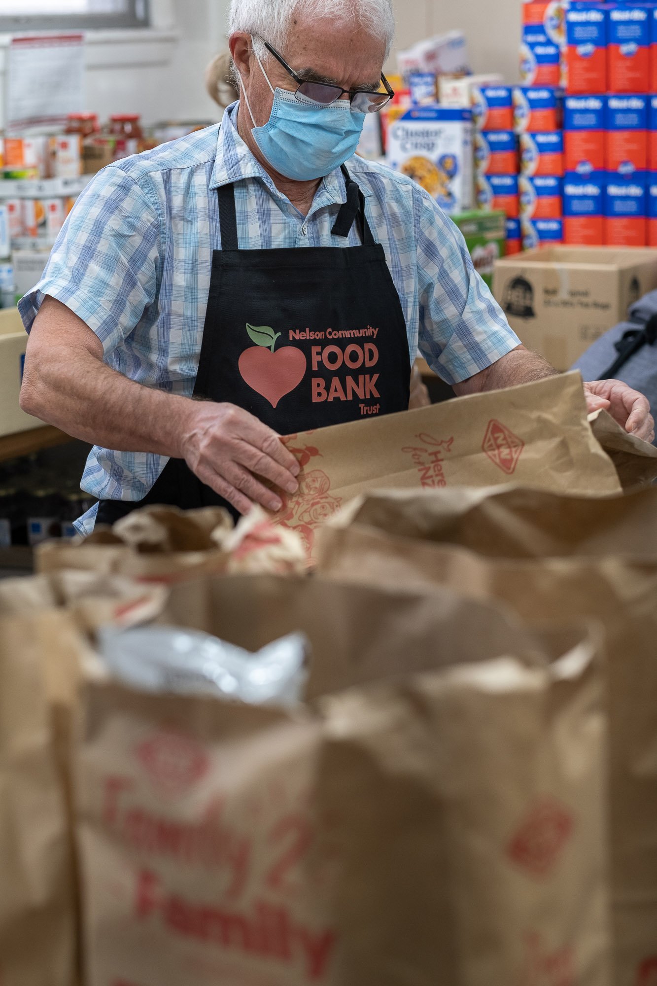 An elderly man wearing glasses, a face mask, a plaid shirt, and an apron with 'Nelson Community Food Bank Trust' on it, sorting through food donations in a food bank.