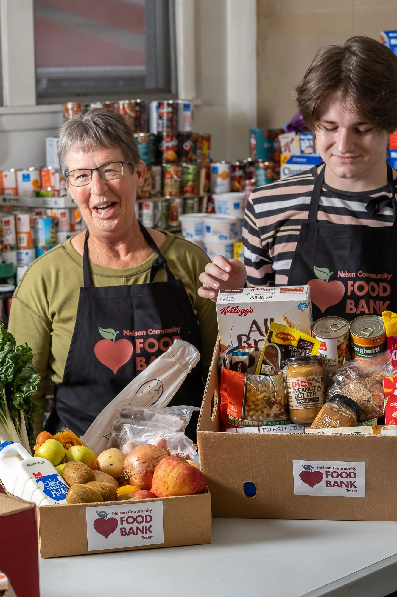 Two people wearing black apron with Nelson Community Food Bank logo working at a food bank, placing grocery items including fruits, vegetables, and canned goods on a table.