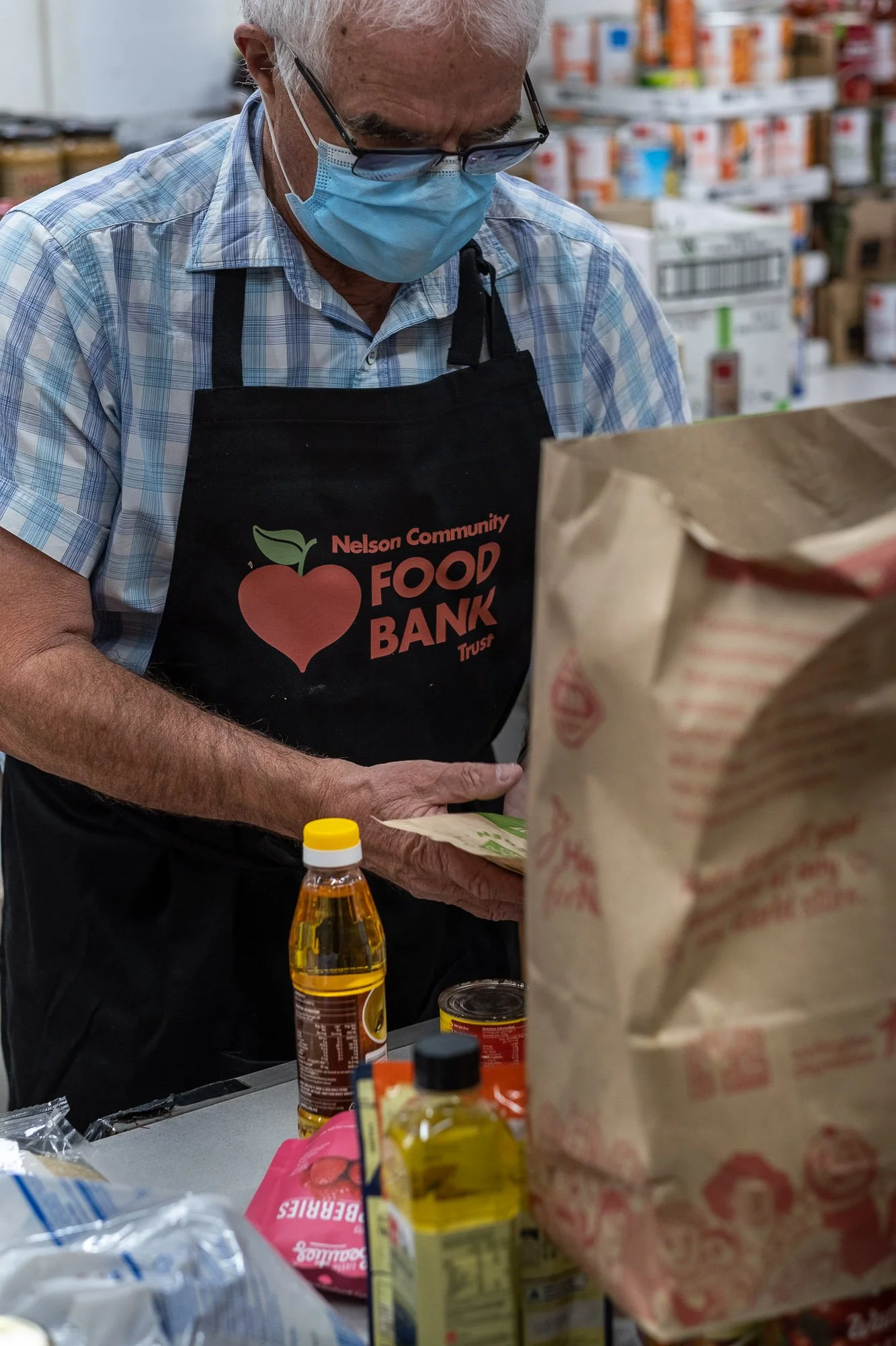 An elderly man wearing glasses and a blue face mask, dressed in a plaid shirt, is shopping at a food bank. He is wearing a black apron that says 'Nelson Community Food Bank Trust' with a red heart and green leaf logo. The table in front of him has bo