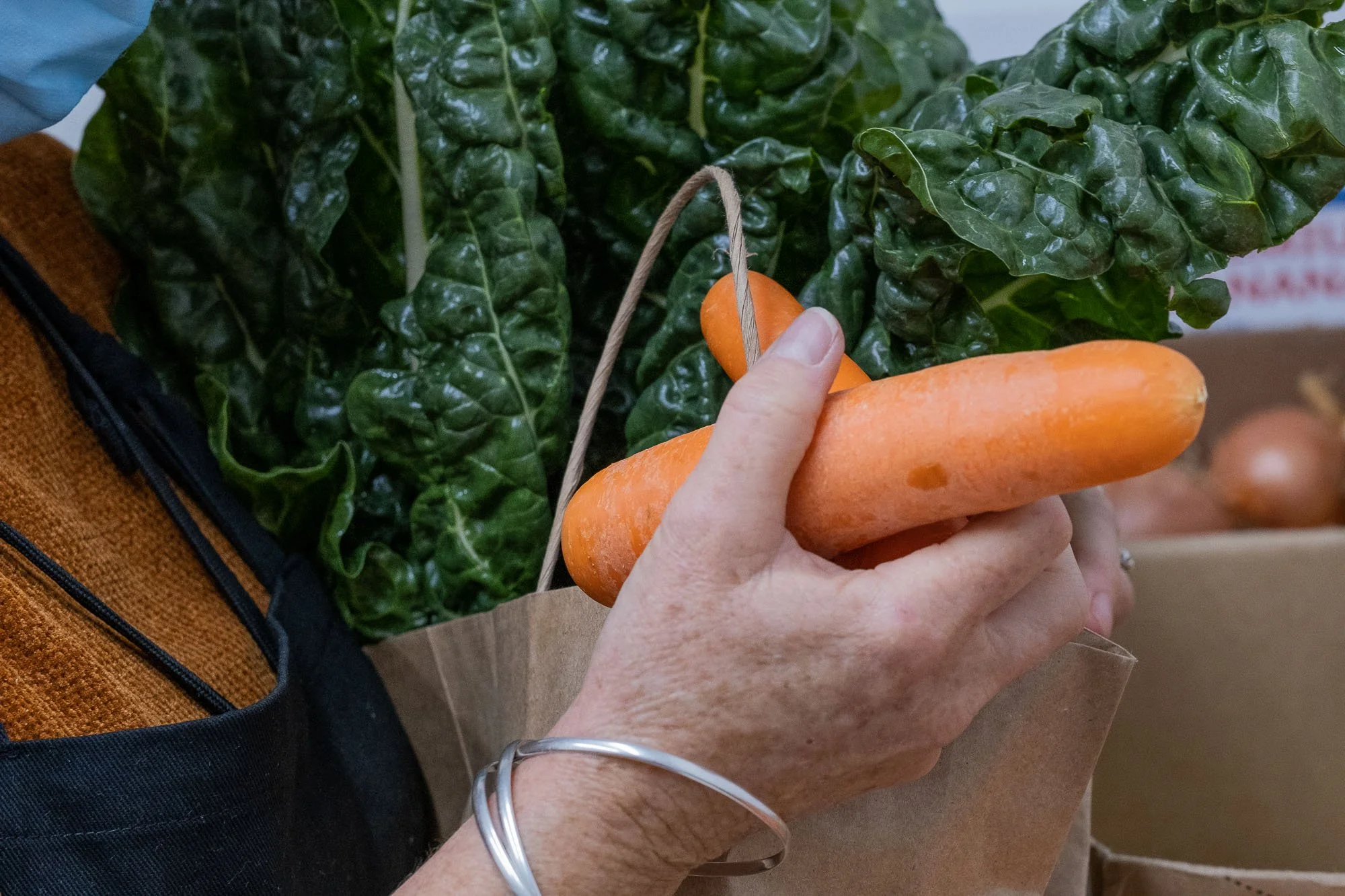 A person holding a carrot in front of a bunch of green leafy vegetables, possibly Swiss chard, in a grocery store.