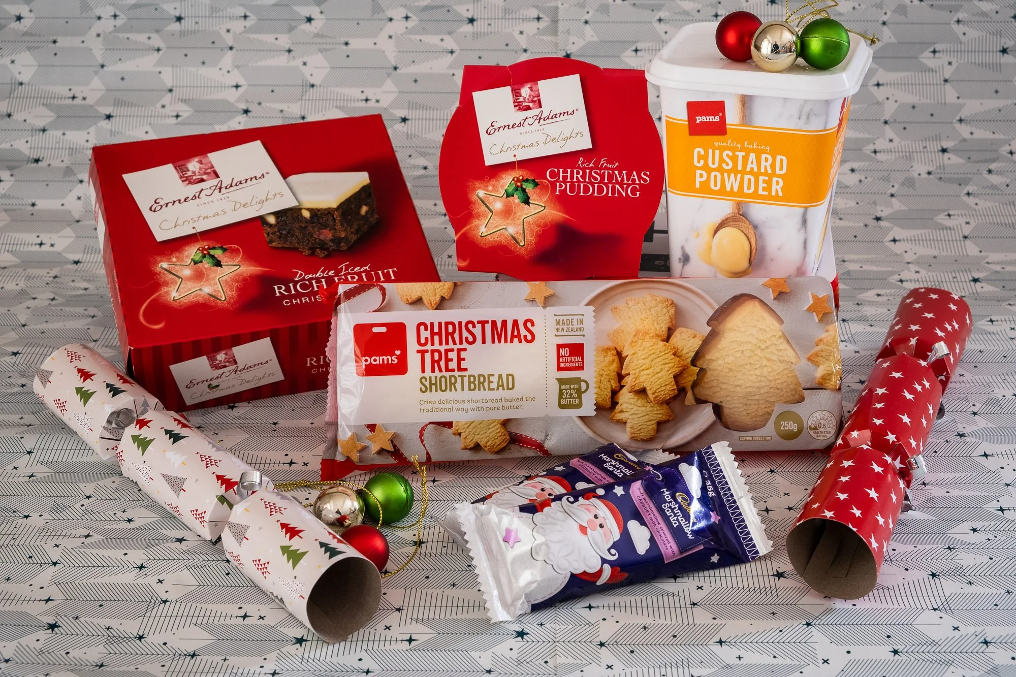 Christmas-themed baked goods, shortbread cookies shaped like trees and stars, Christmas pudding, double iced fruit cake, custard powder in a container, Christmas crackers with festive patterns, Christmas ornaments, and marshmallows with Santa Claus d
