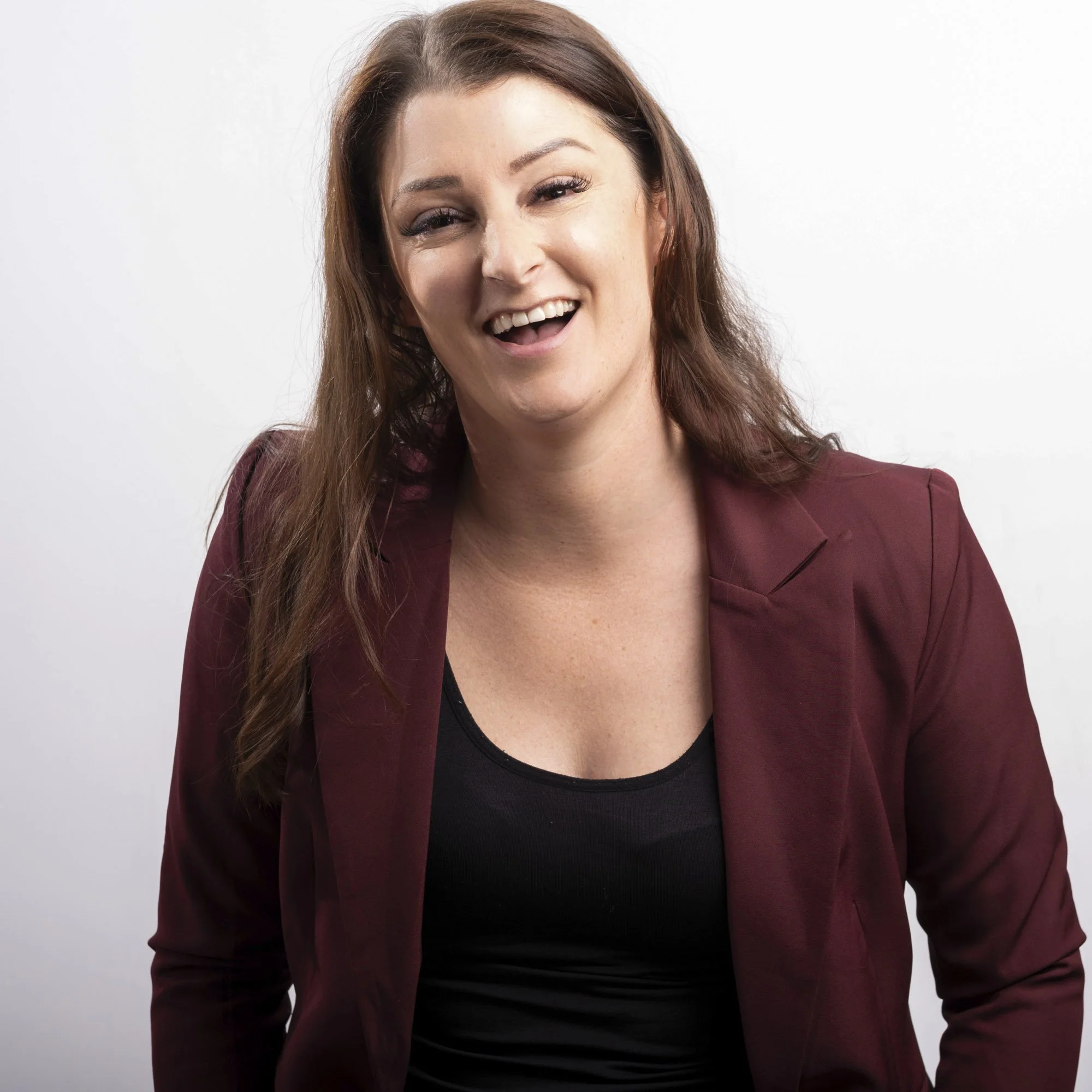 A woman with long, wavy brown hair smiling and winking in front of a plain white background, wearing a dark top and a maroon blazer.