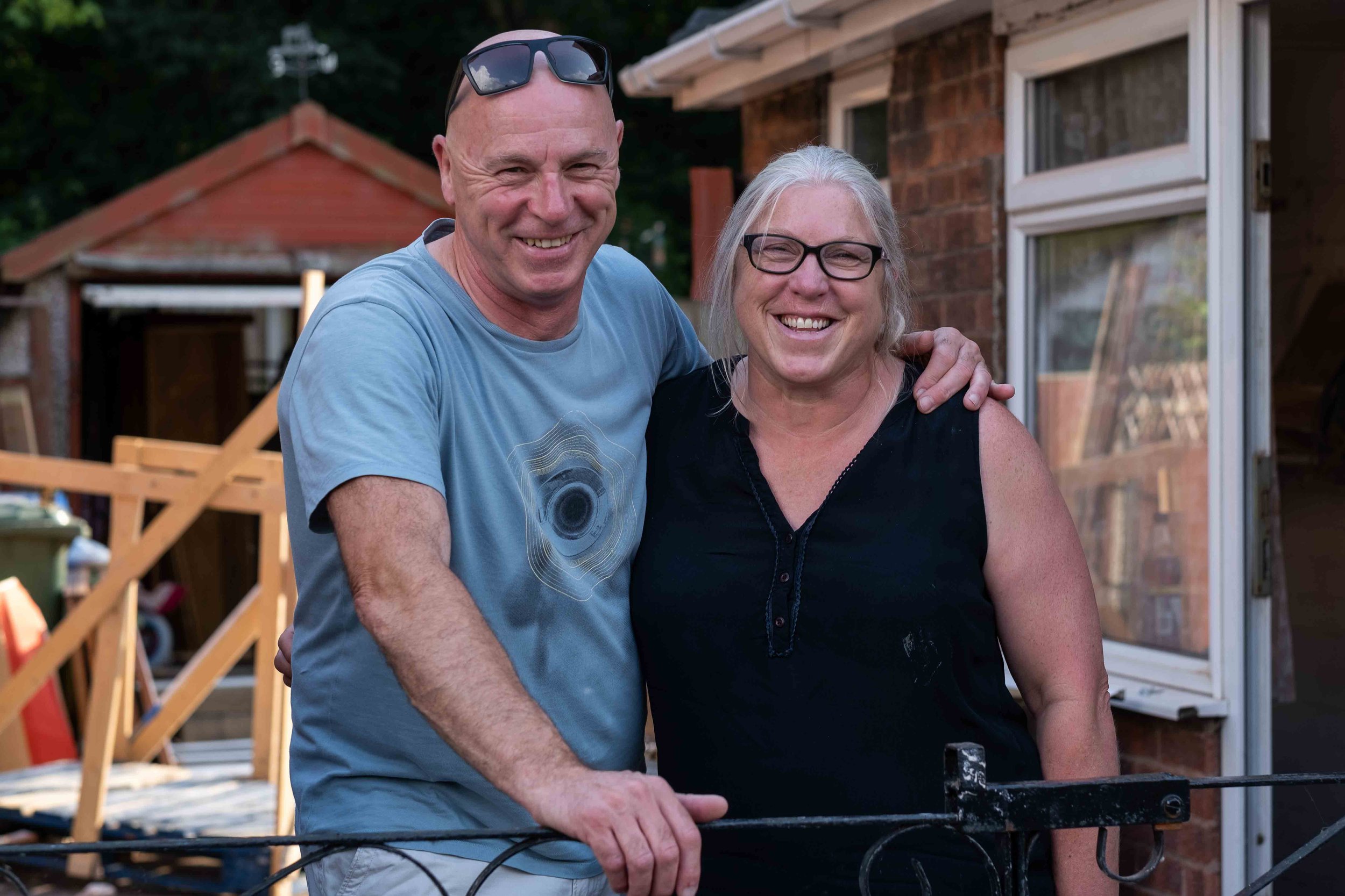 A happy couple smiling and standing outdoors in front of a brick house. The man is wearing a blue t-shirt and sunglasses on his head, and the woman is wearing a black sleeveless top and glasses. They have their arms around each other.