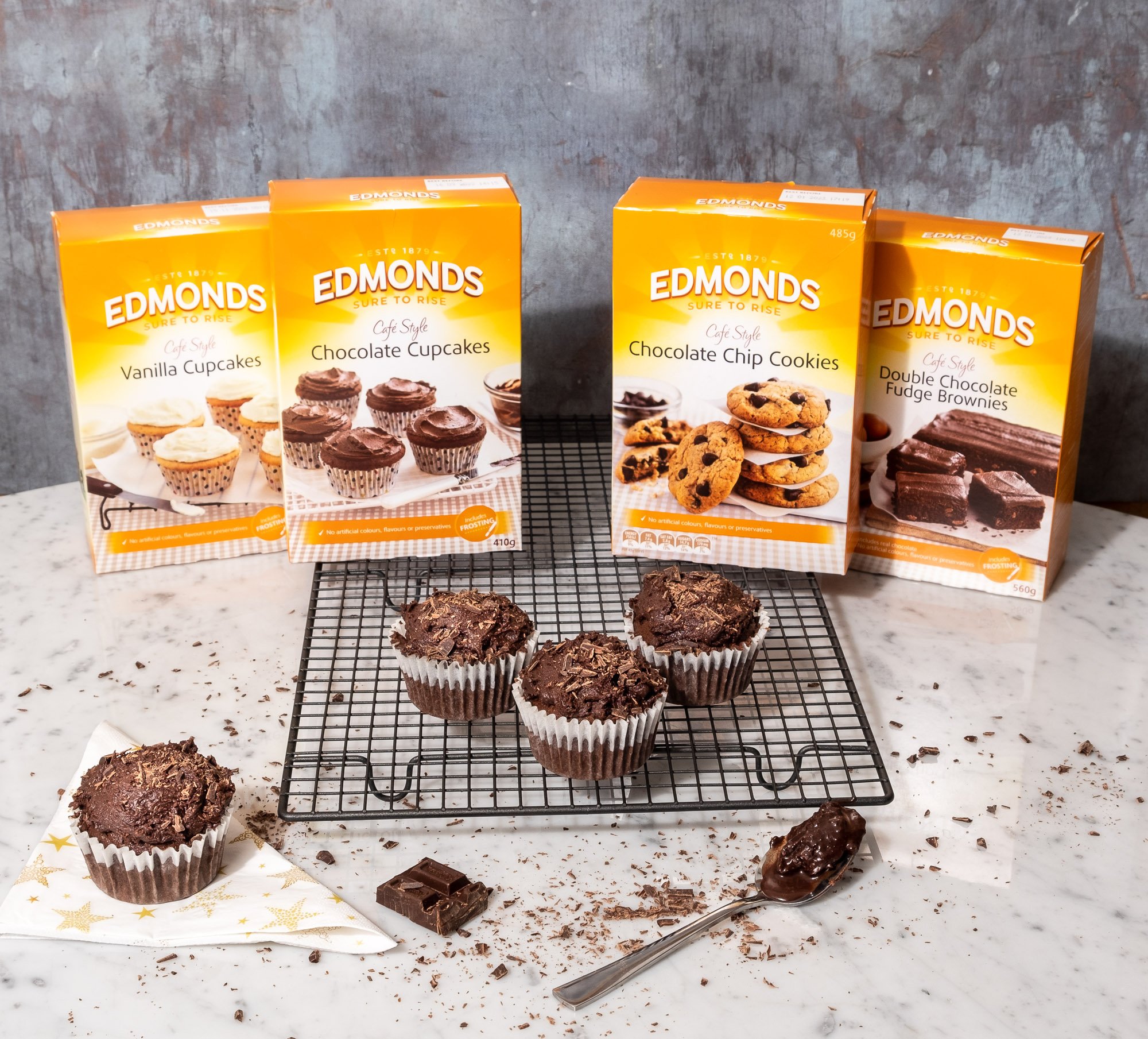 An assortment of semi-finished chocolate muffins on a wire rack surrounded by chocolate pieces, a spoon with chocolate, and boxes of Edmonds baking ingredients including vanilla cupcakes, chocolate cupcakes, chocolate chip cookies, and double chocola