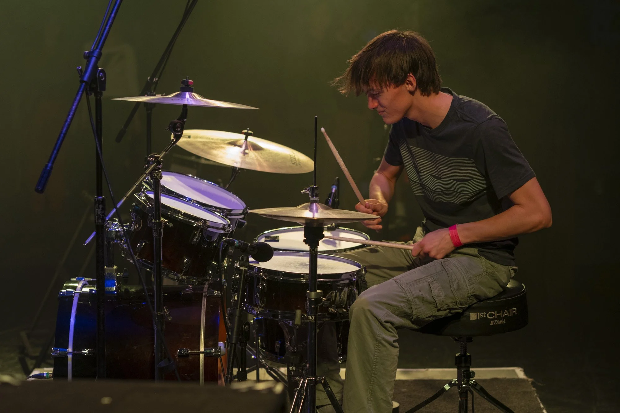 A person with brown hair playing a drum set on stage. They are seated on a black drum throne and holding drumsticks, with a focused expression. A microphone is positioned near the drums, and the background is dark.