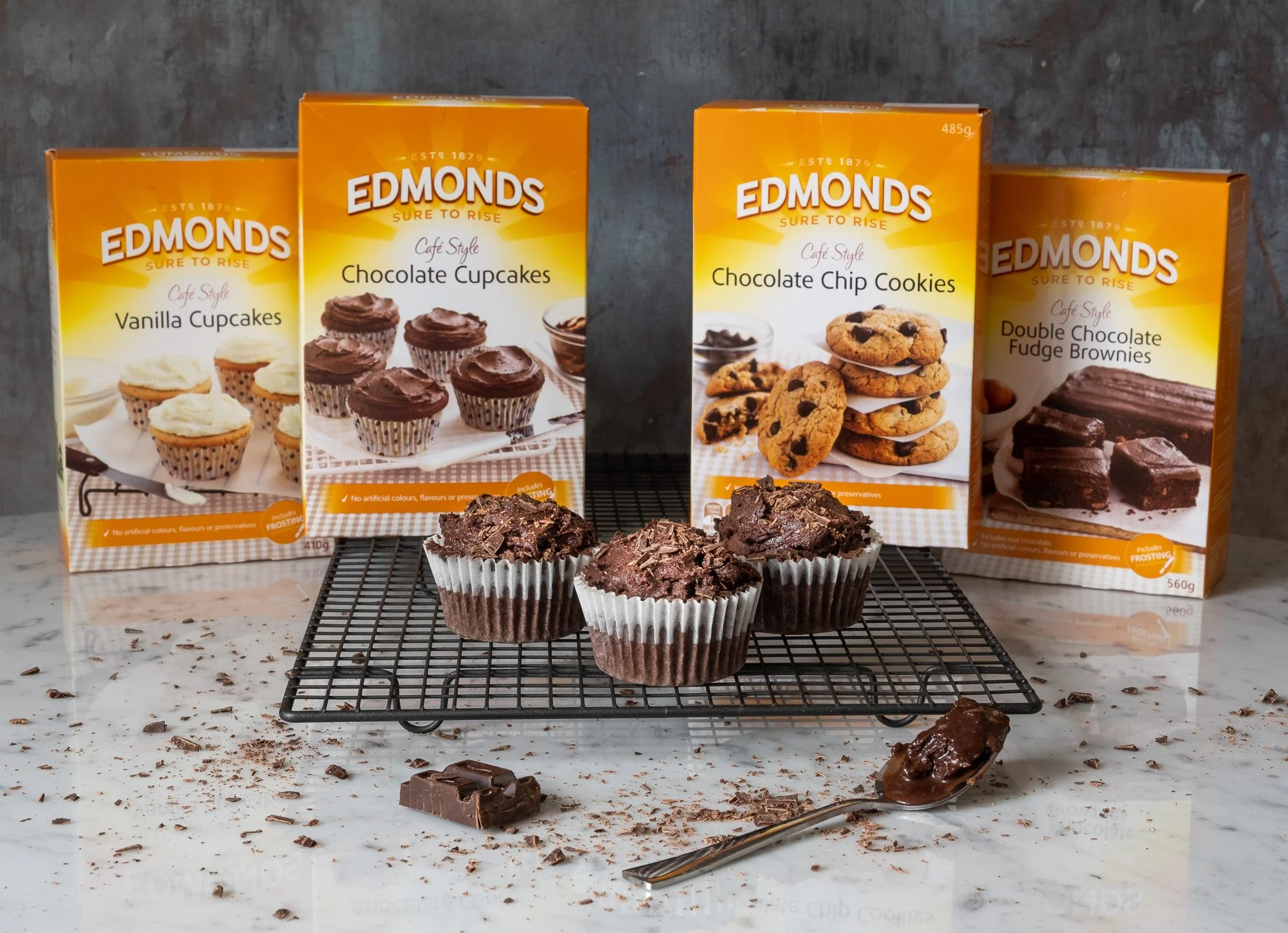Three chocolate cupcakes on a cooling rack, surrounded by four Edmonds baking mix boxes for vanilla cupcakes, chocolate cupcakes, chocolate chip cookies, and chocolate fudge brownies, with chocolate pieces and a spoon of chocolate on a marble counter