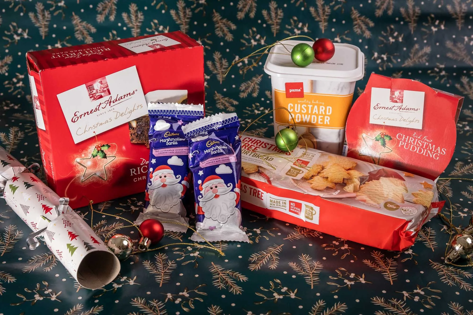 Christmas-themed assortment of snacks and decorations, including boxes of Ernest Adams Christmas delights, marshmallow Santa candies, a container of custard powder, a box of Christmas pudding, a partially unrolled holiday wrapping paper, Christmas or