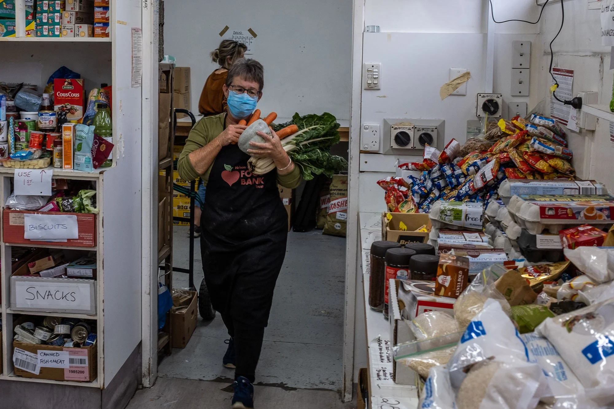 A volunteer wearing a face mask and apron carries vegetables inside a food pantry, with shelves stocked with canned goods, snacks, and other food items.