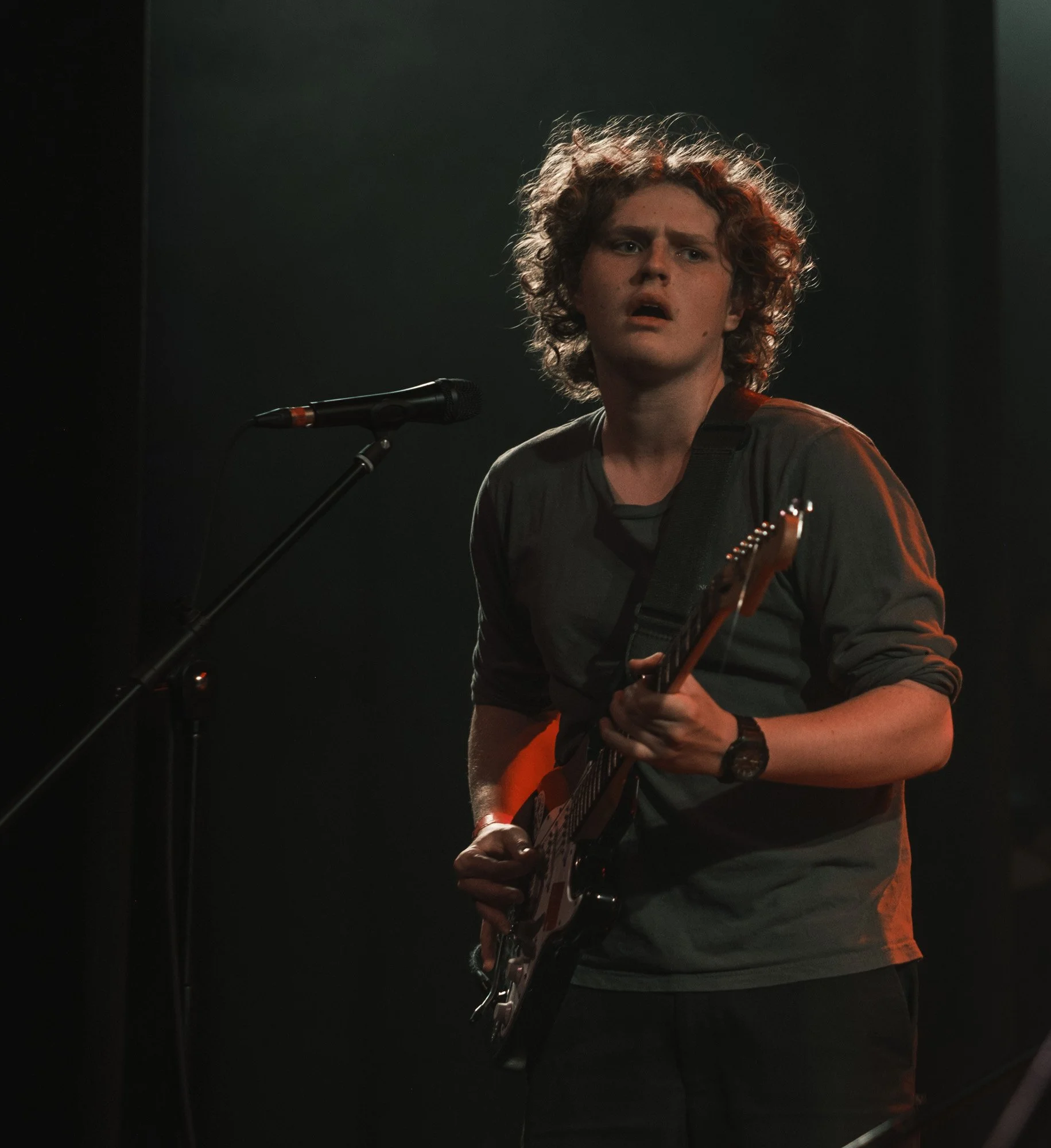 Young man with curly hair playing an electric guitar and singing into a microphone on stage, wearing a dark t-shirt and watch, with stage lights creating a dark background.
