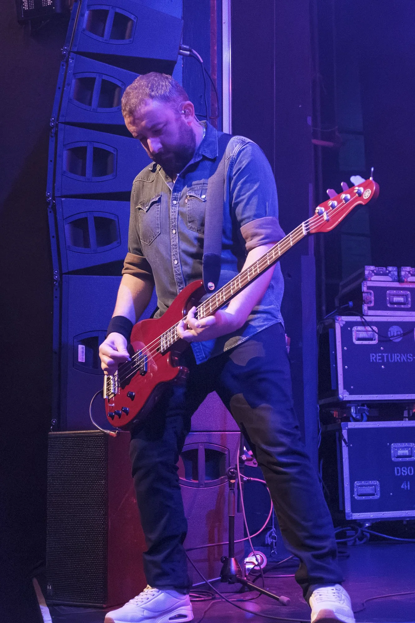 A musician playing a red electric guitar on a stage with black walls and equipment including speakers and amplifiers, under purple stage lighting.