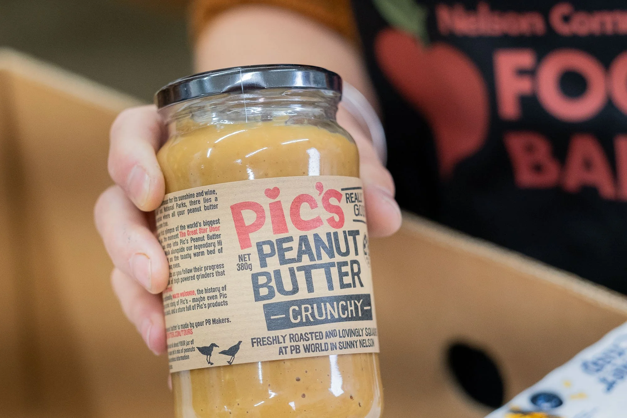 A hand holding a jar of Pic's Peanut Butter Crunchy. The label reads 'Pic's Peanut Butter - Crunchy' and indicates it is freshly roasted and lovingly squished at PB World in Sunny Nelson.