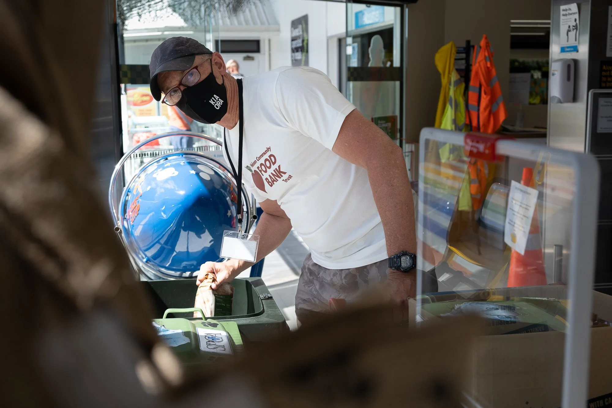 A man wearing a black face mask, glasses, a white Food Bank T-shirt, and a cap is placing a bag into a bin at a food bank. He is inside a store or donation center with safety vests, cones, and signs visible in the background.