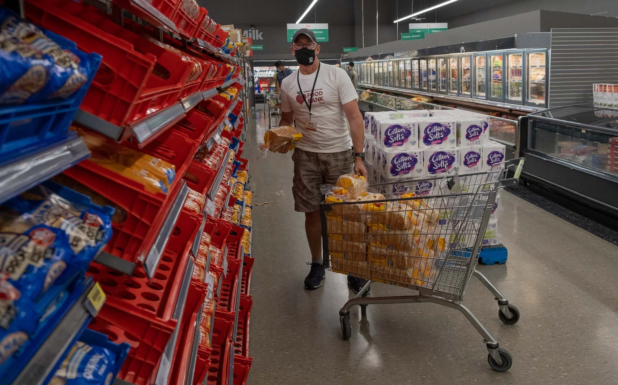 A man shopping at a grocery store, wearing a face mask, white t-shirt, and camouflage shorts, pushing a cart filled with bread and other items, walking down an aisle with snacks and frozen goods.