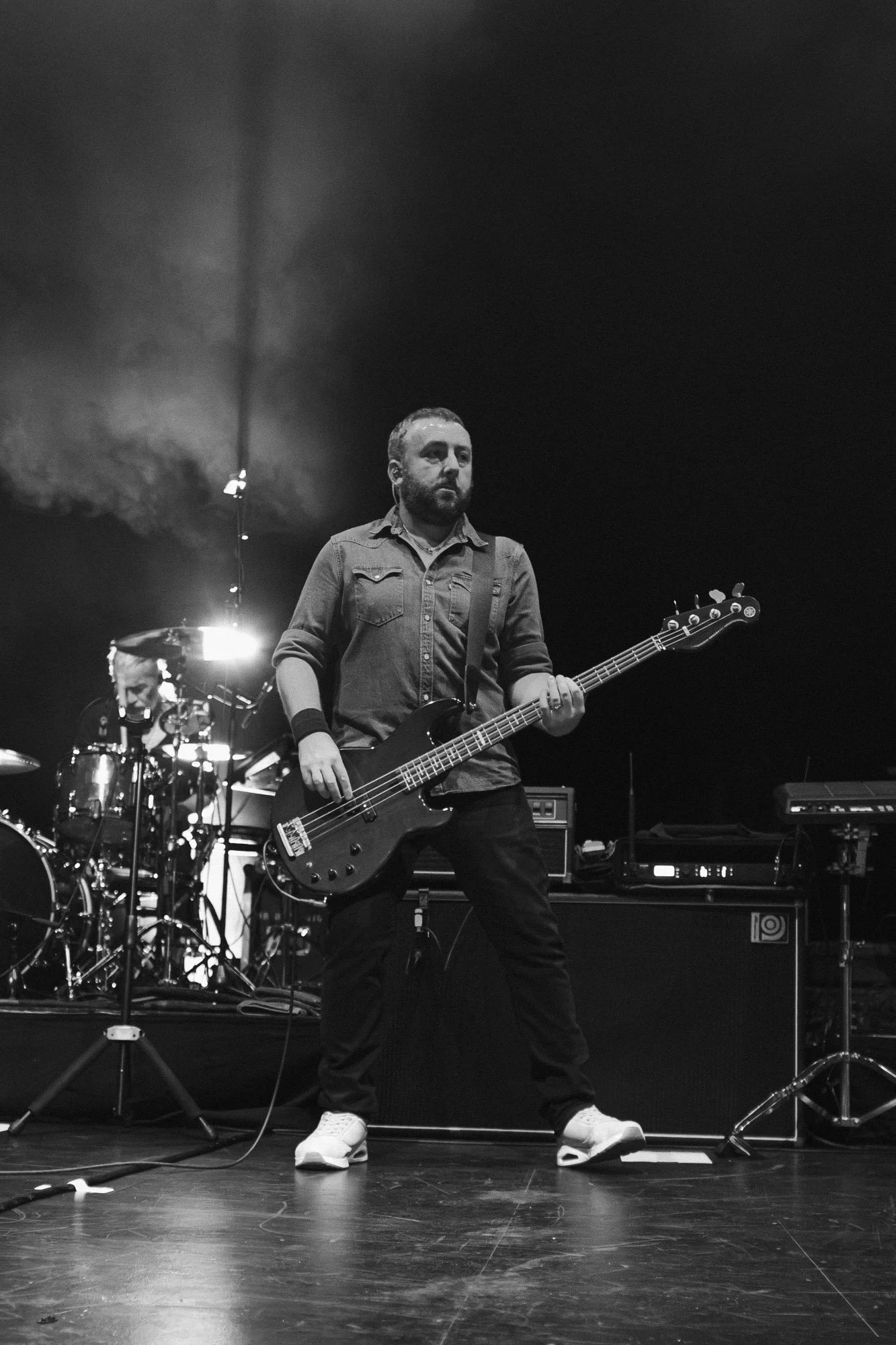 Black and white photo of a man playing a bass guitar on stage, with a drummer and musical equipment in the background.