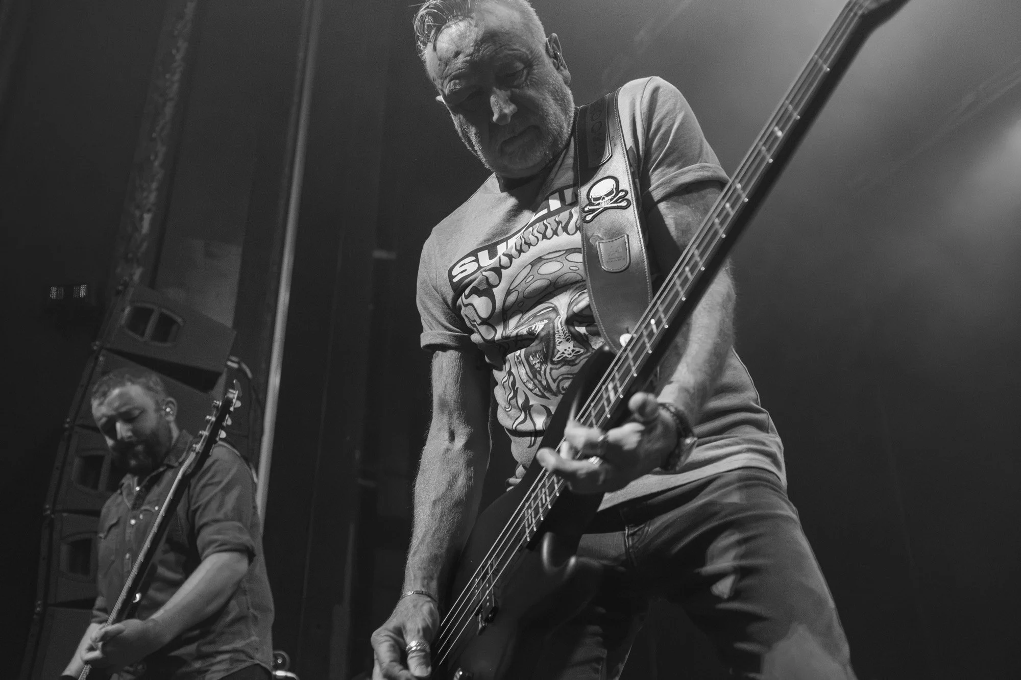 Two male musicians passionately playing electric guitars on stage during a live performance, with a focus on the musician in the foreground wearing a t-shirt with graphic prints.