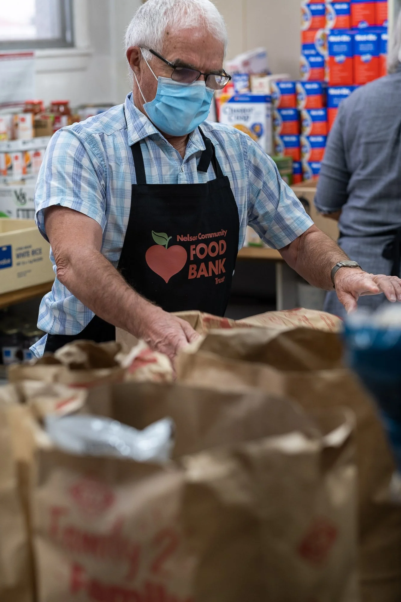 An elderly man wearing glasses, a face mask, and a Nelson Community Food Bank apron is packing food in a donation center.