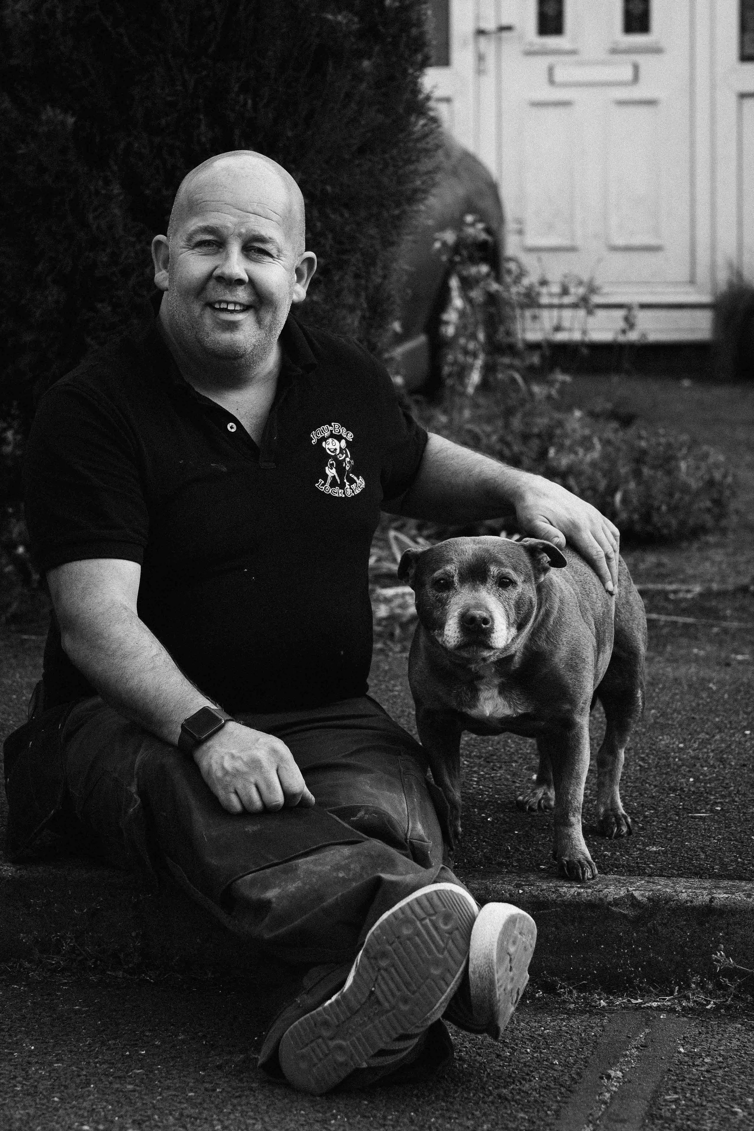 A man kneels on the ground next to a dog in an outdoor setting. The man is smiling and wearing a black T-shirt with a logo. The dog is a mixed breed with a sturdy build, standing next to him.