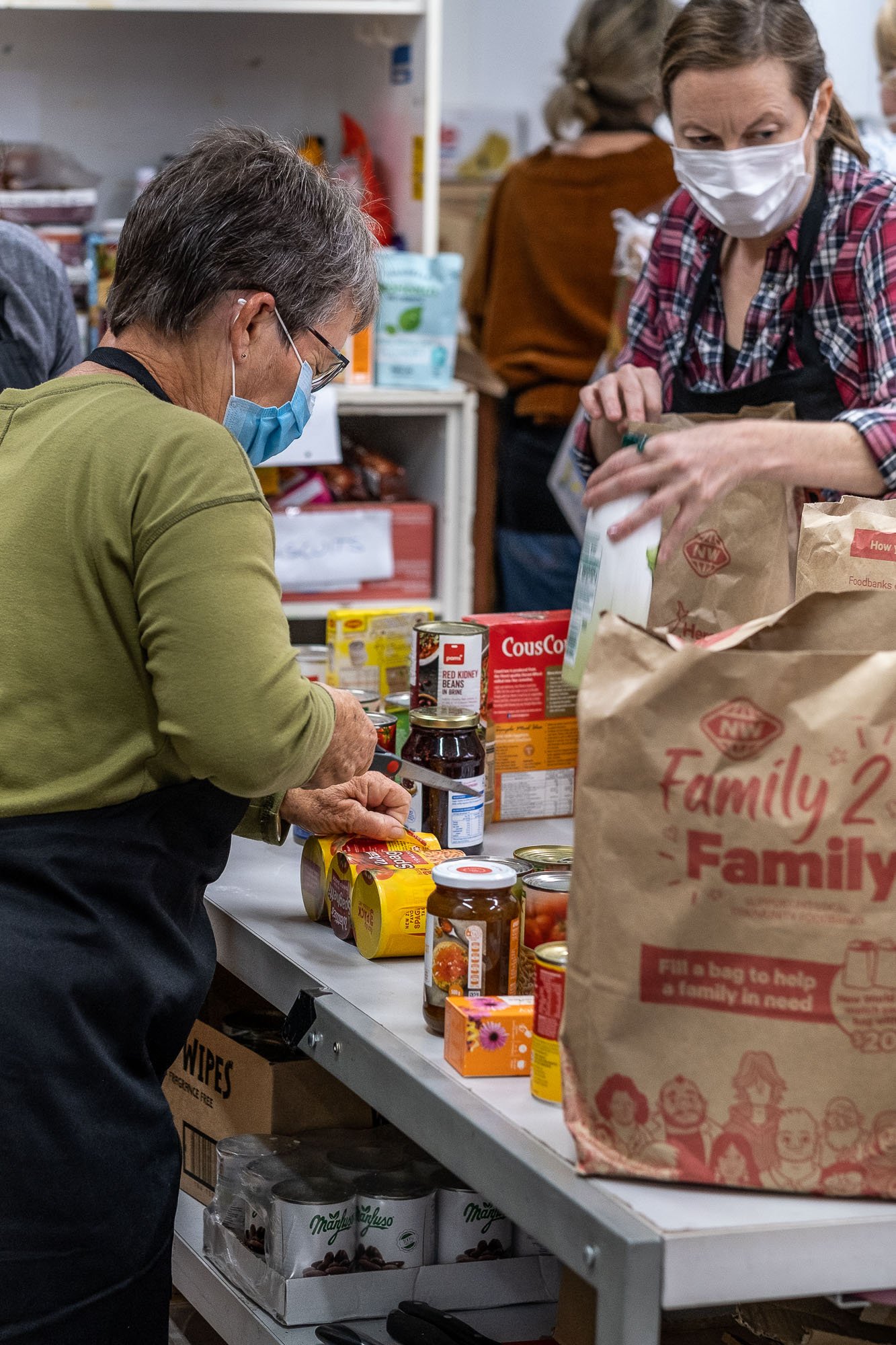 Two women, both wearing masks, sorting food donations at a community food bank. One woman in a green shirt is selecting items from a table filled with canned goods and boxed foods, while the other woman in a red plaid shirt looks on.