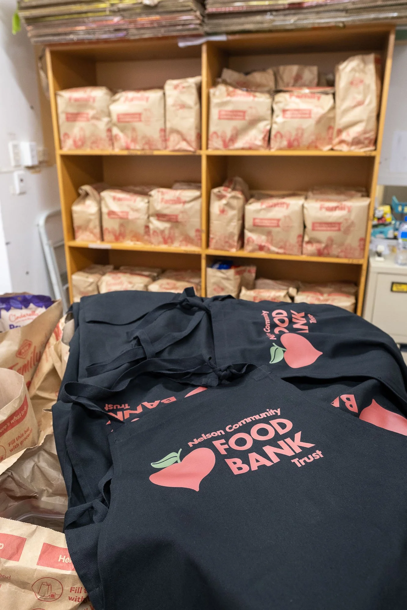 Black tote bags with Nelson Community Food Bank Trust logo placed on a table in front of a wooden shelf filled with paper bags of donated food.