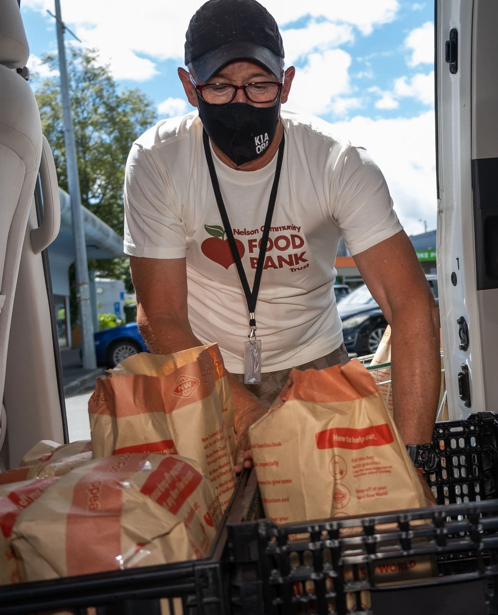 A man wearing a black face mask, glasses, and a white t-shirt with the logo of the Nelson Community Food Bank, loads paper grocery bags into a vehicle, outdoors with a partly cloudy sky.