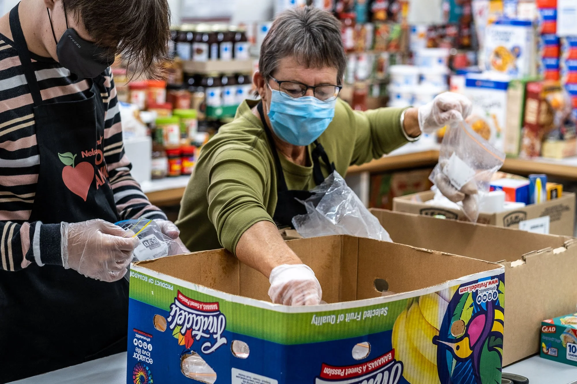Two women wearing masks and gloves packing food into a box at a food bank or pantry, with boxes and canned goods in the background.