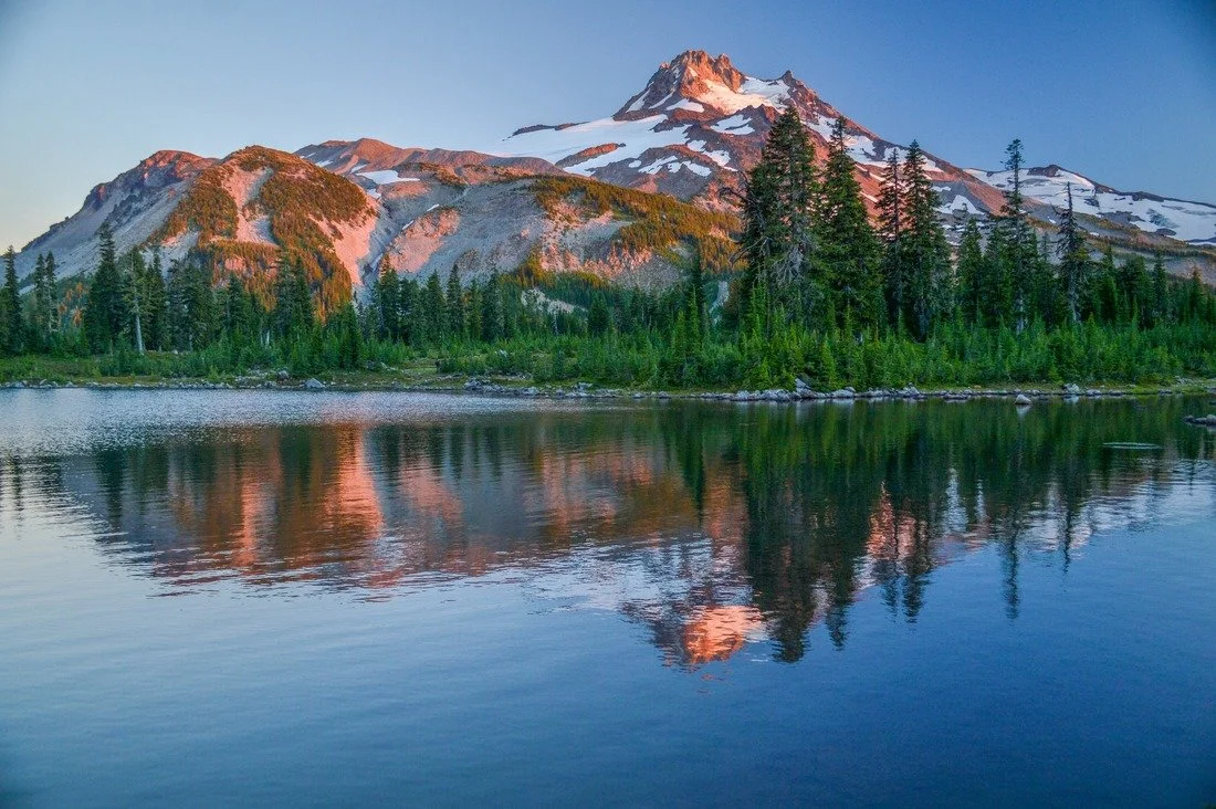 A mountain with snow-capped peaks and green forest reflected in a lake at dusk.