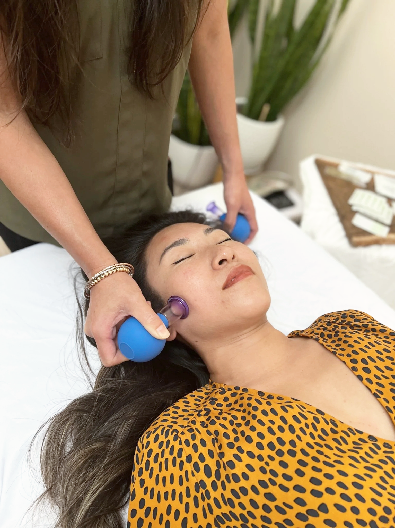 A woman lying on a treatment bed with her eyes closed while receiving a facial or skincare treatment from a practitioner using facial cupping