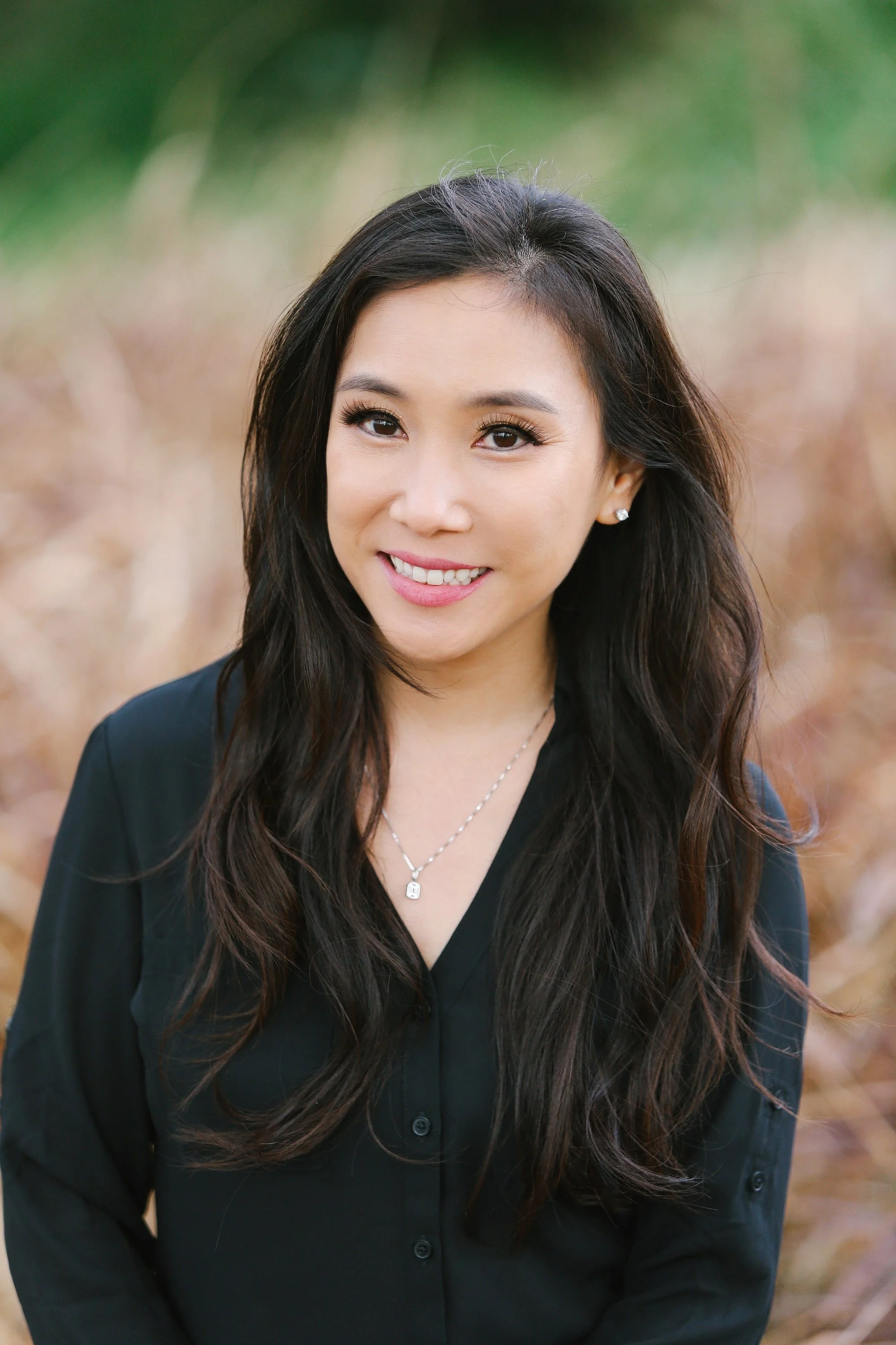 A woman with long dark hair smiling outdoors, wearing makeup, a black top, jewelry, and earrings with a blurred natural background.