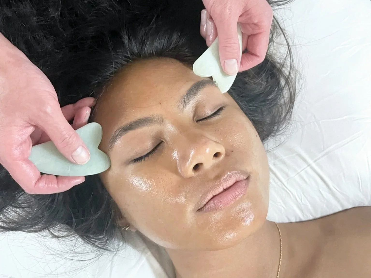 A woman receiving a facial treatment with jade rollers on her forehead, lying down with her eyes closed.