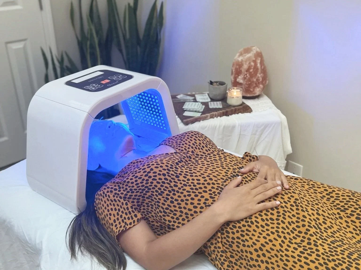 A woman receiving a blue light therapy treatment in a therapy device while lying on a bed, with a table in the background holding salt lamp and candles.