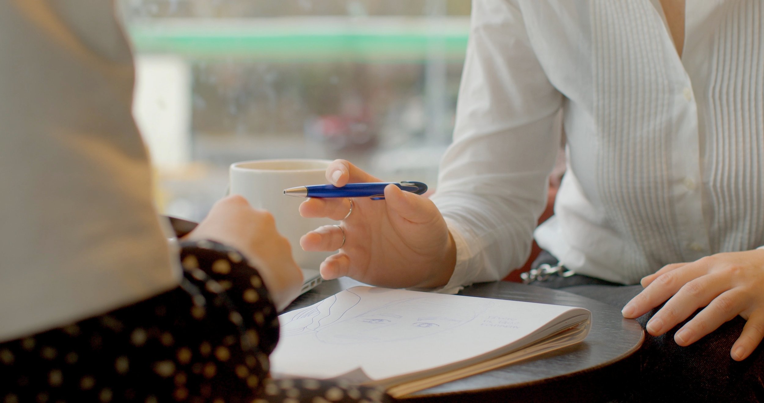 People sitting at a table in a cafe or office, exchanging documents, with one person holding a pen and a white mug.