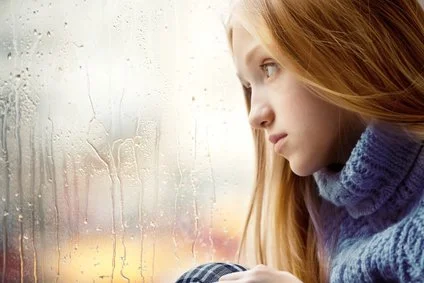 A young woman with red hair gazes out a rainy window, appearing thoughtful or sad.