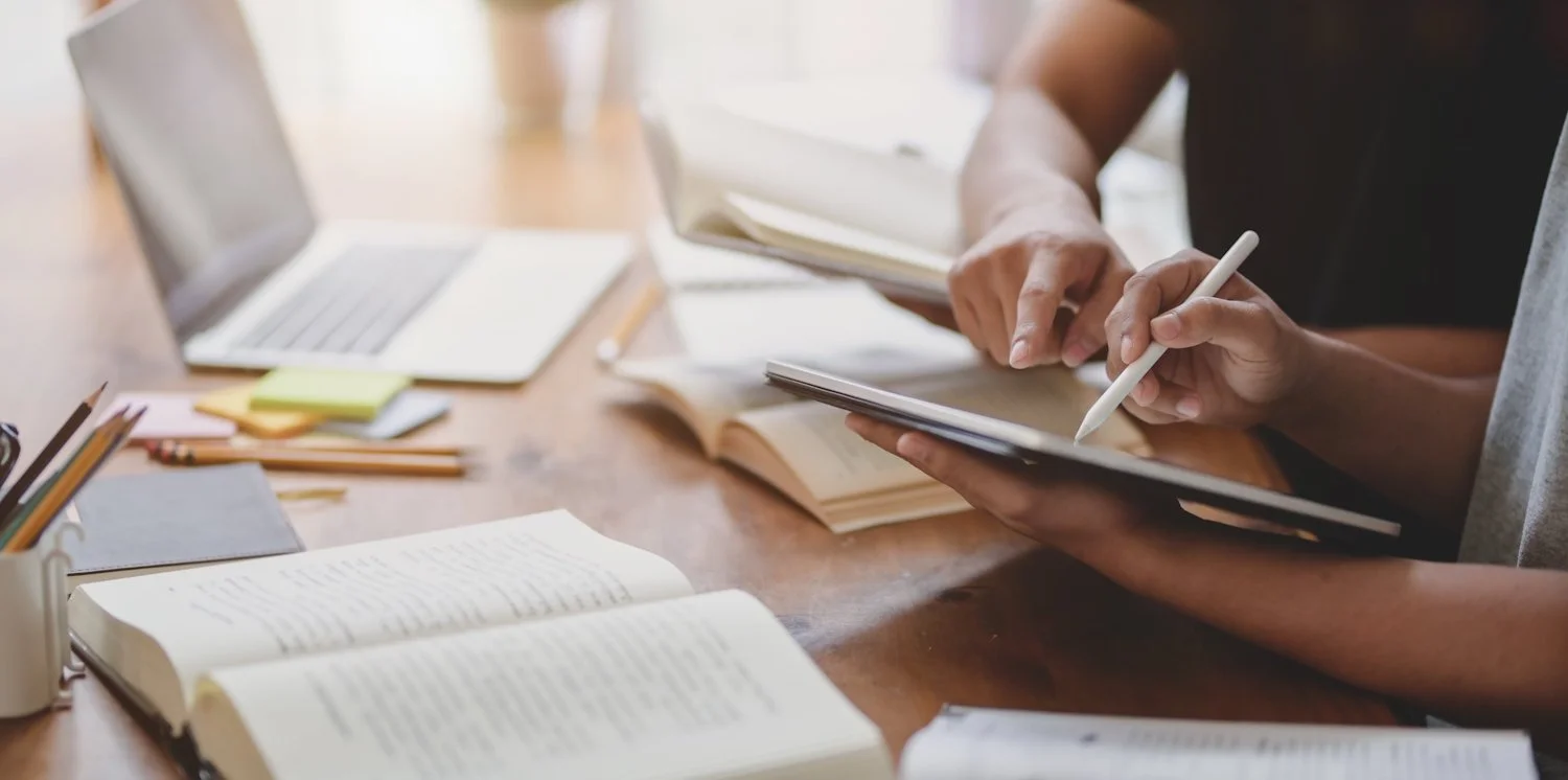 Two people working at a wooden table with open books, notebooks, a laptop, and writing utensils.