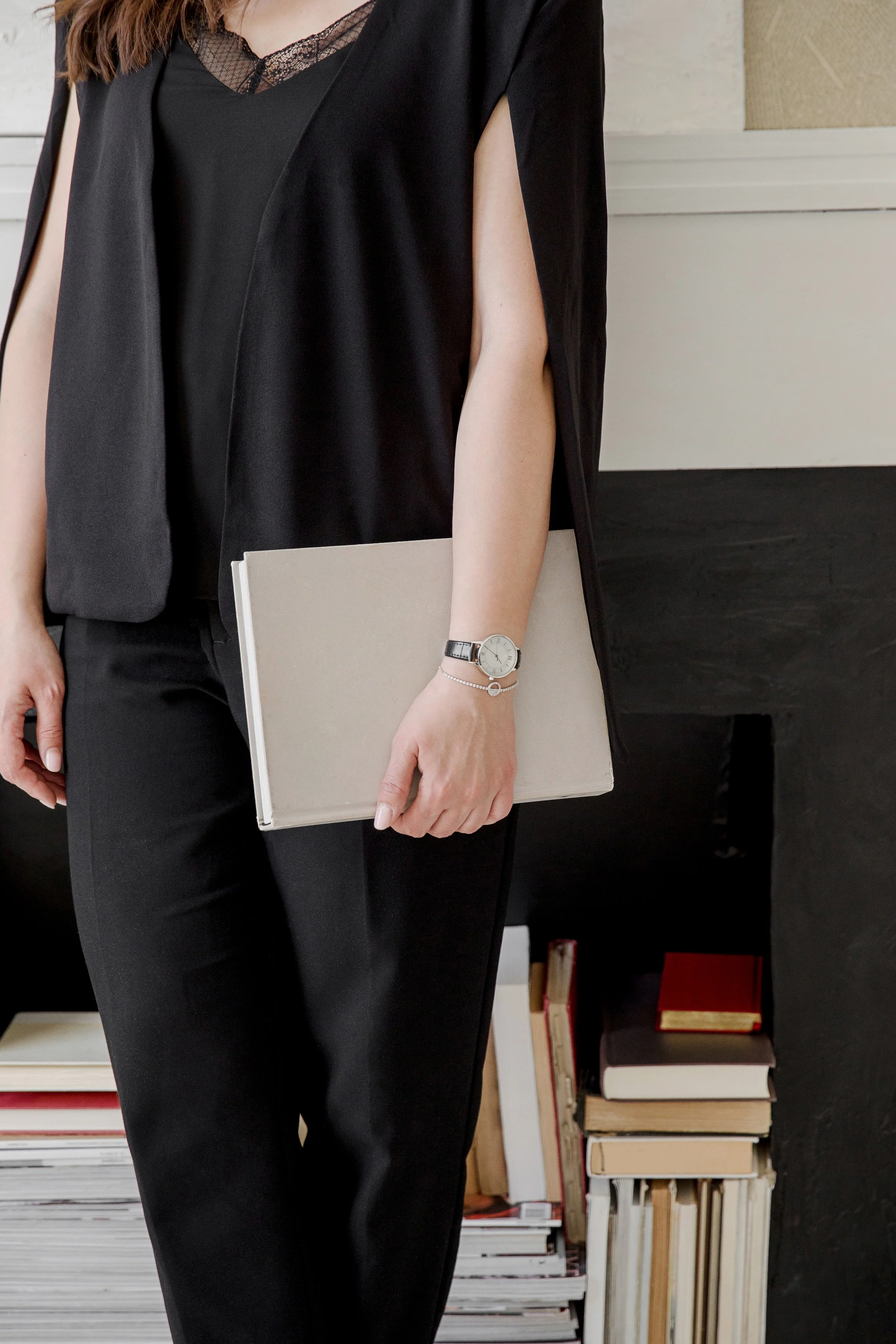 A woman dressed in black yoga pants and a black top, wearing a silver watch and bracelet, holding a closed beige notebook, standing in front of a bookshelf filled with books.