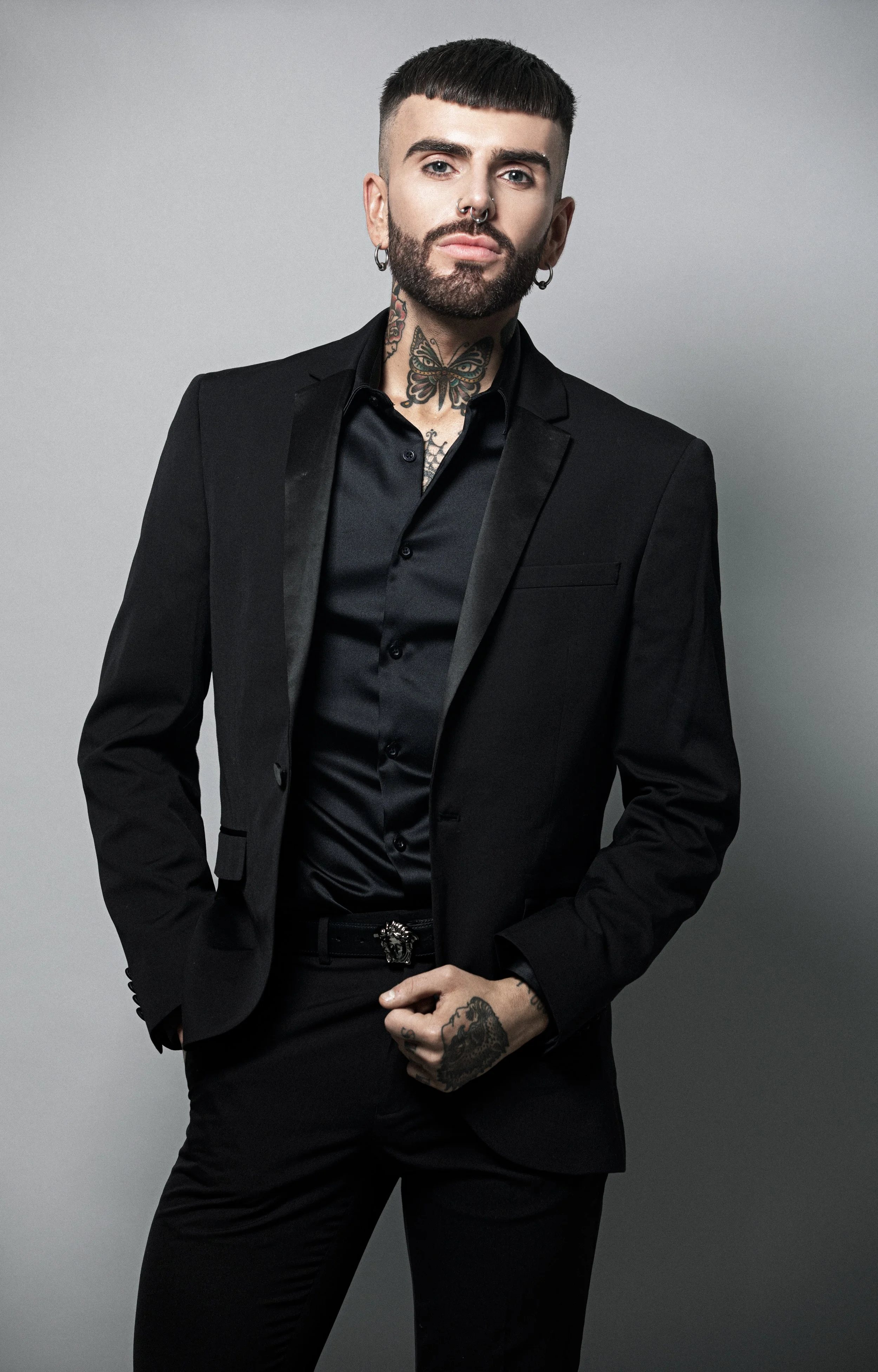 A man with a beard and tattoos wearing a black suit and shirt, posing against a plain gray background.