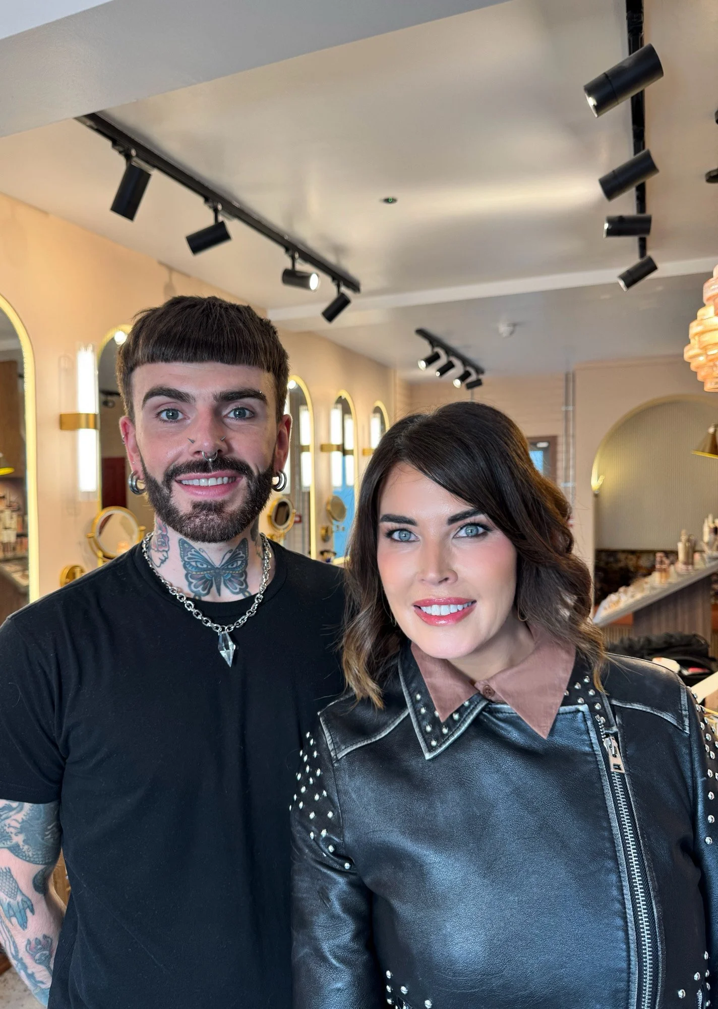 A man with tattoos, piercings, and a woman with shoulder-length wavy hair posing inside a well-lit room with arched mirrors and stylish decor.