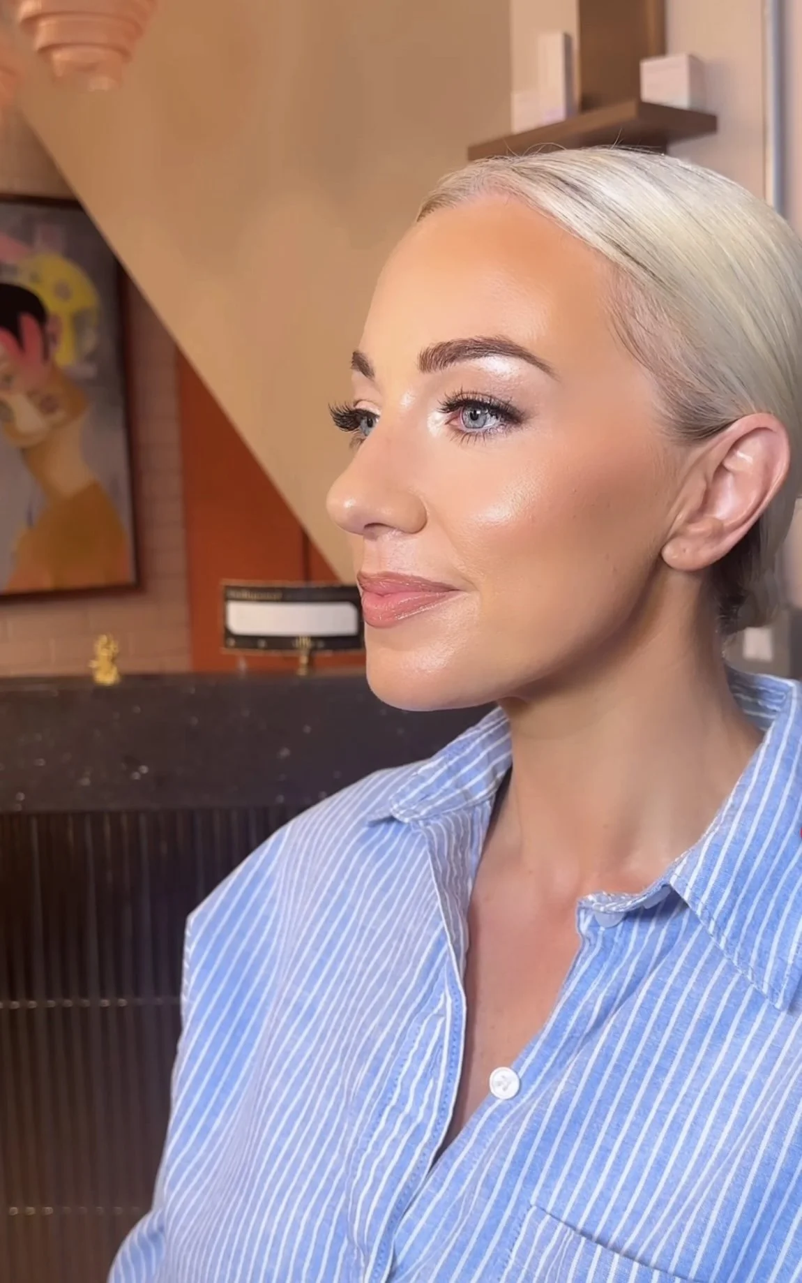 A woman with blonde hair, blue eyes, and makeup, wearing a blue and white striped shirt, is indoors with artwork on the wall behind her.