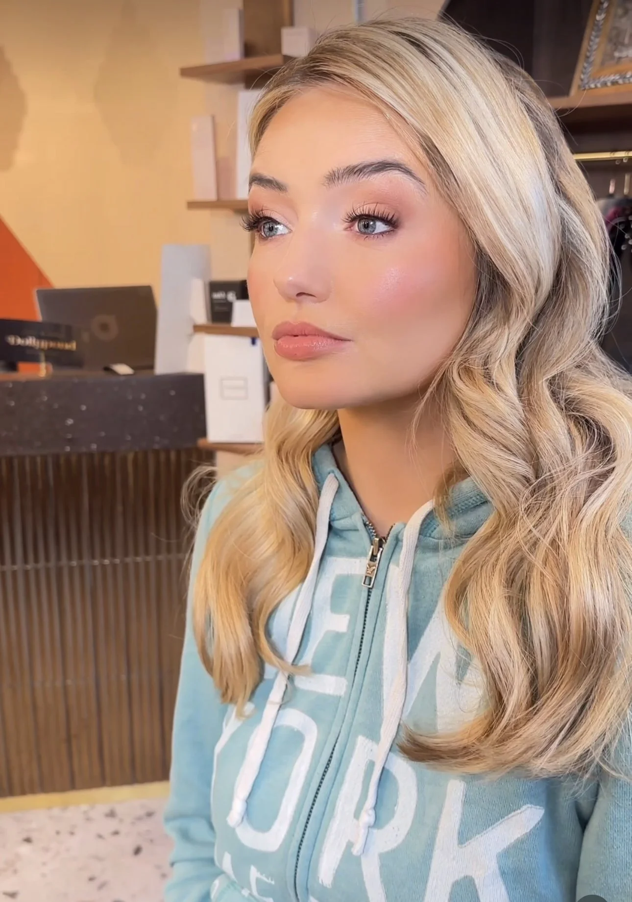 Close-up of a young woman with long blonde wavy hair, wearing a blue hoodie, in an indoor setting.