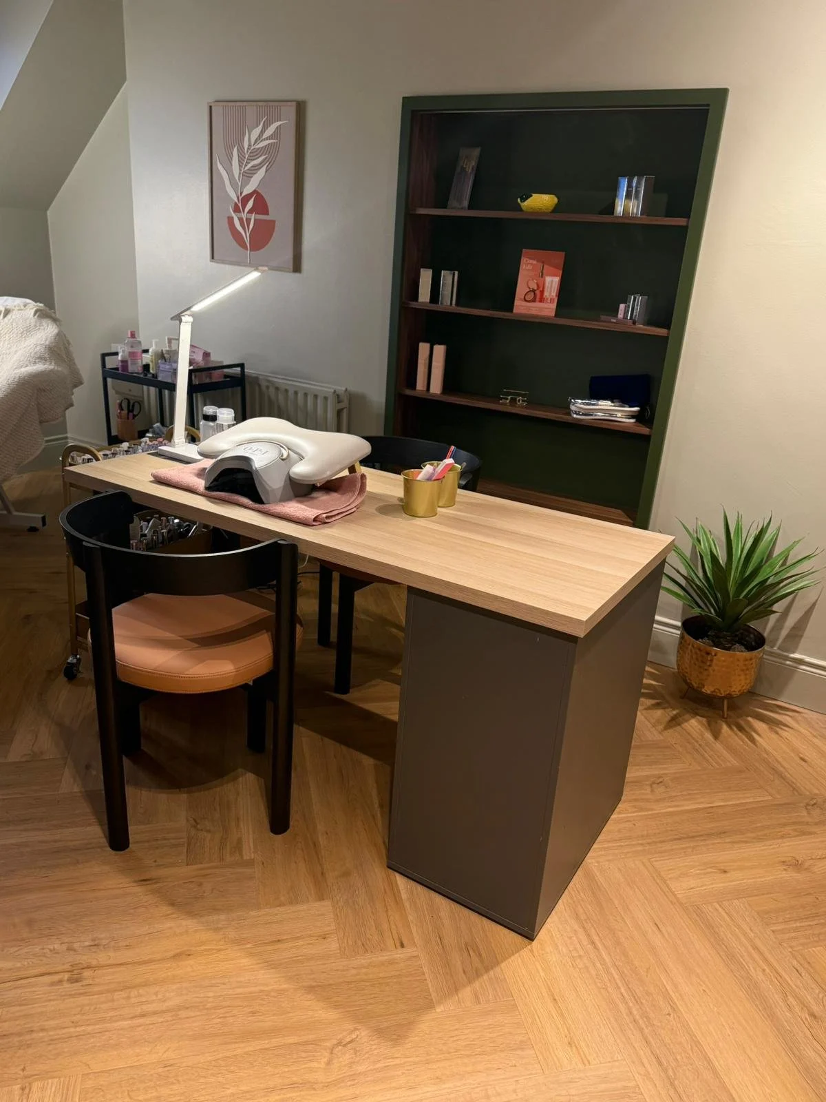 A manicure or beauty salon workspace with a wooden table, a chair with a peach cushion, and a green bookshelf in the background. The table has a nail lamp and various manicure tools, with a small gold container holding pink and white pens. A potted plant is on the floor to the right, and there are decorative items and books on the bookshelf.