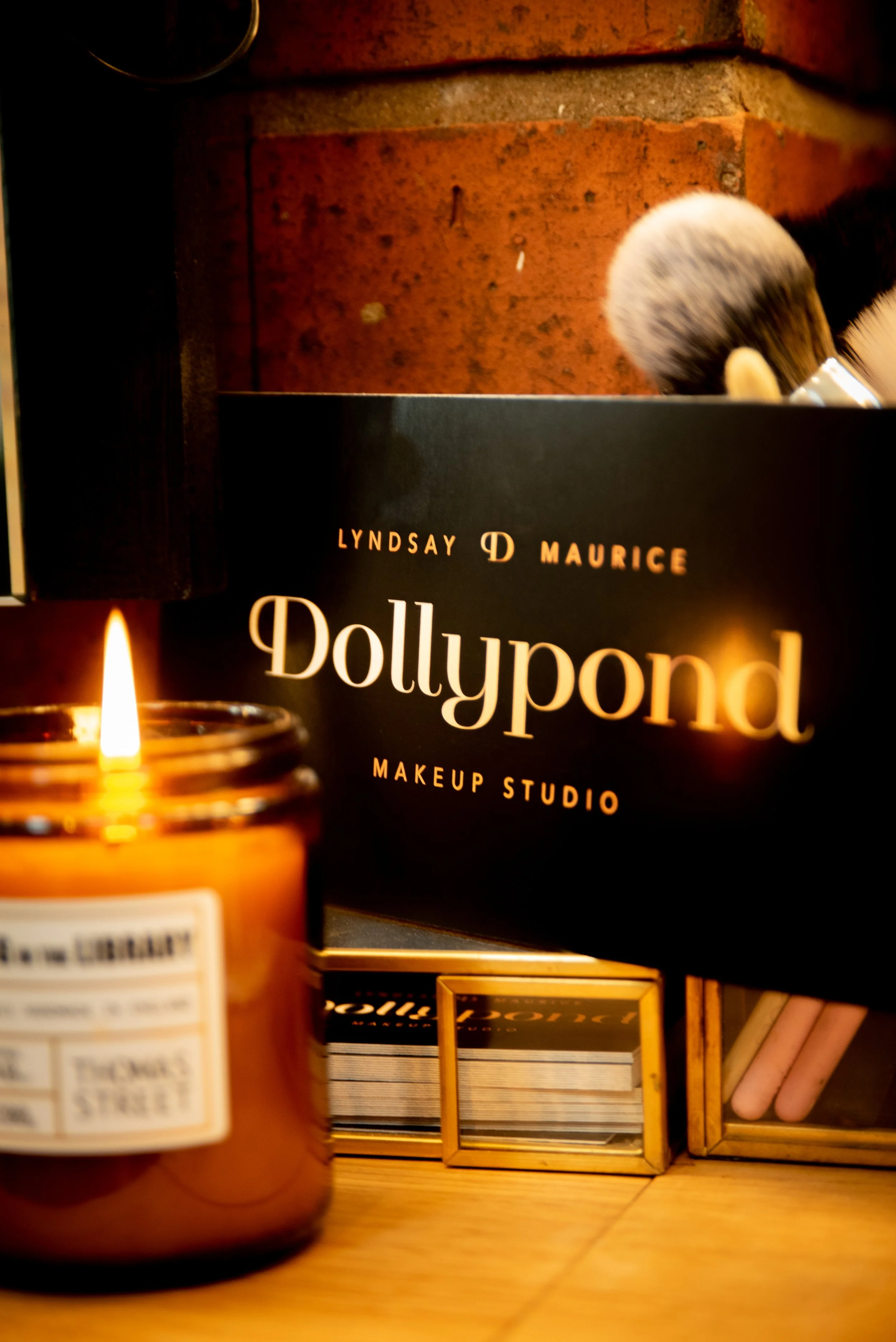 Beauty studio branding box labeled 'Dollypod Makeup Studio,' a lit candle, and small boxes with makeup tools on a wooden surface.