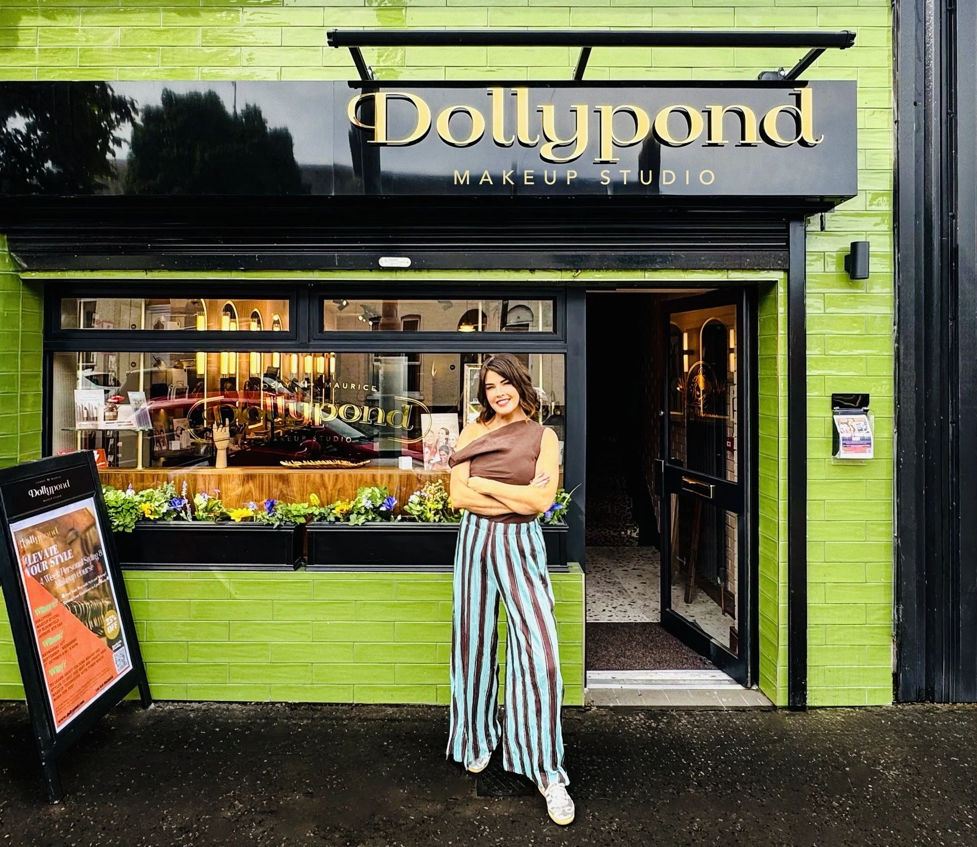 A woman standing outside a makeup studio called Dollypound, smiling, wearing striped pants and a brown top, in front of a green building with a large window displaying the studio's sign and interior.