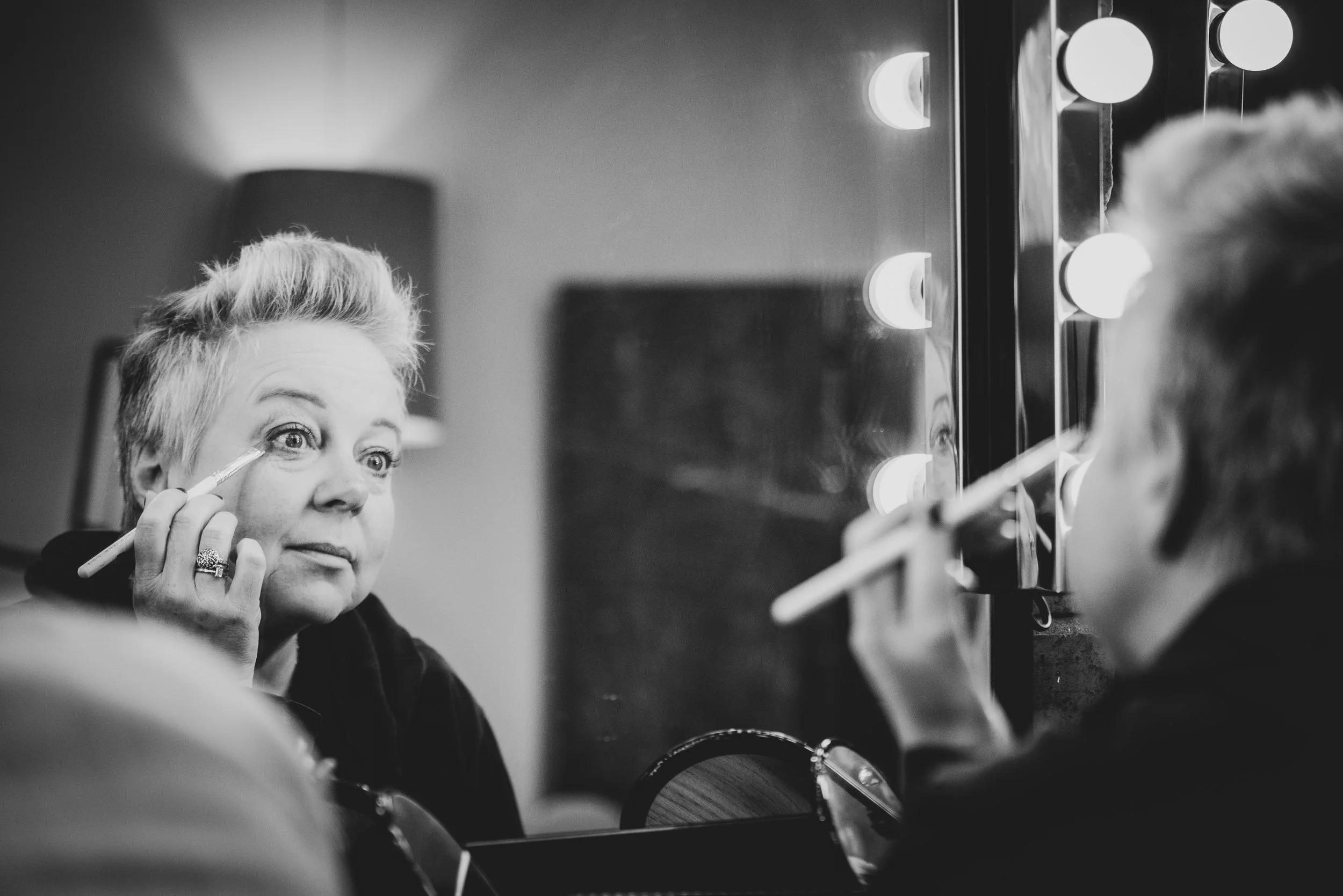 A woman applying makeup while looking in a mirror with light bulbs around it